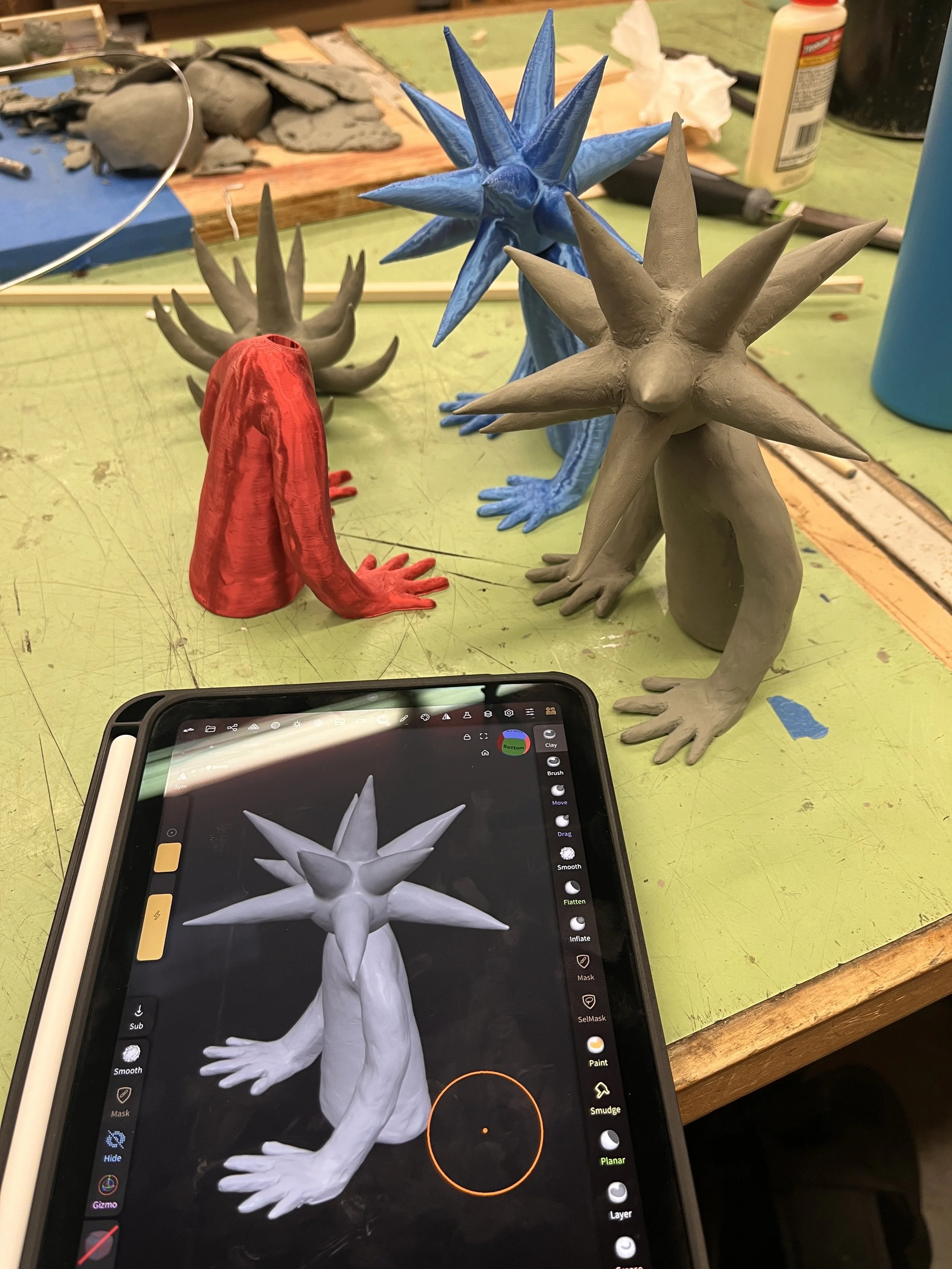 Handmade clay sculptures of various creatures with spiky heads and long limbs, in different colors (gray, red, blue). An tablet on the table displays a 3D model of one of the sculptures, showing a creature with elongated limbs and multiple pointed sp