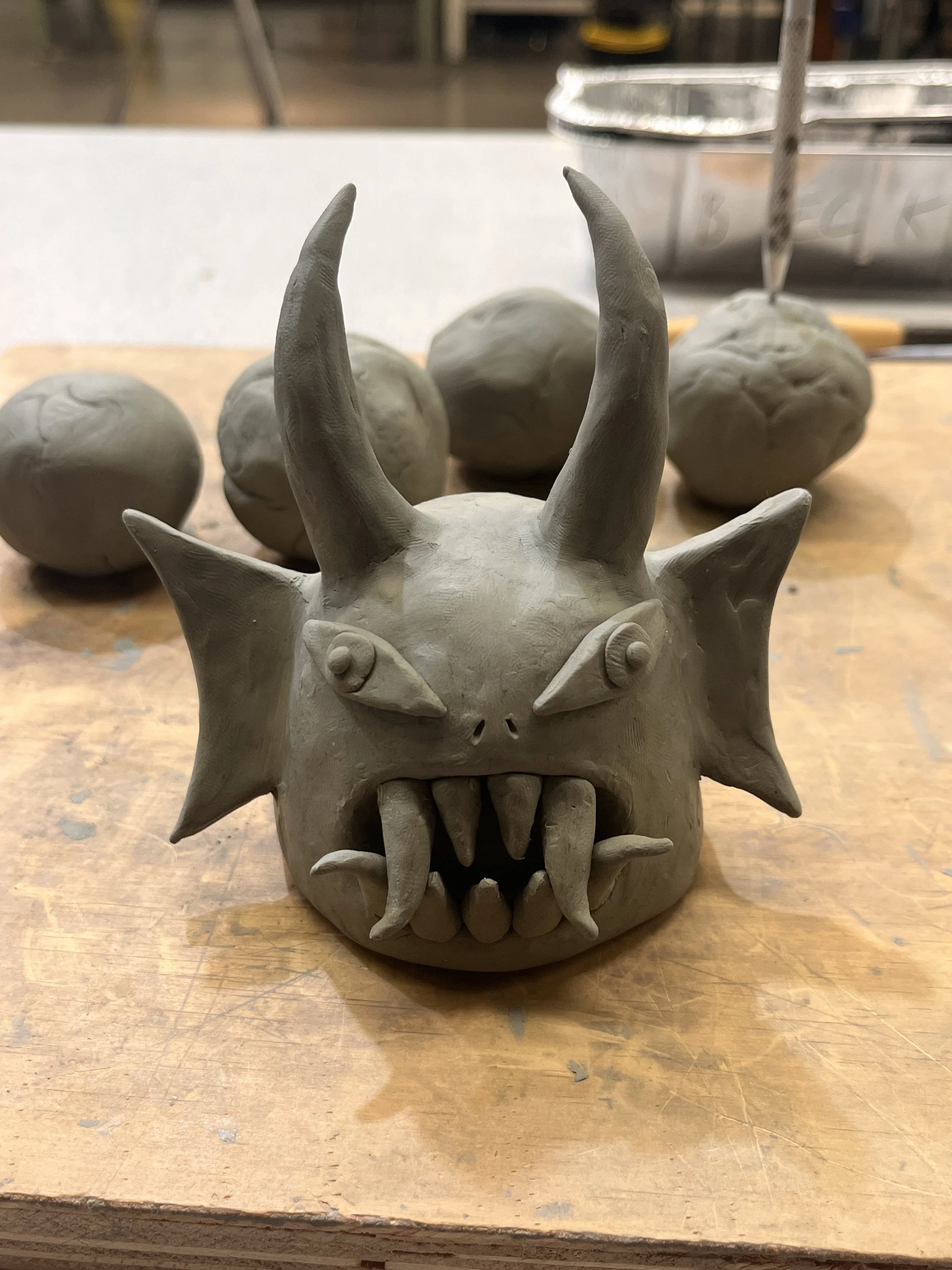 A clay sculpture of a monster with horns, spikes, sharp teeth, and large eyes, on a wooden work surface with clay balls in the background.