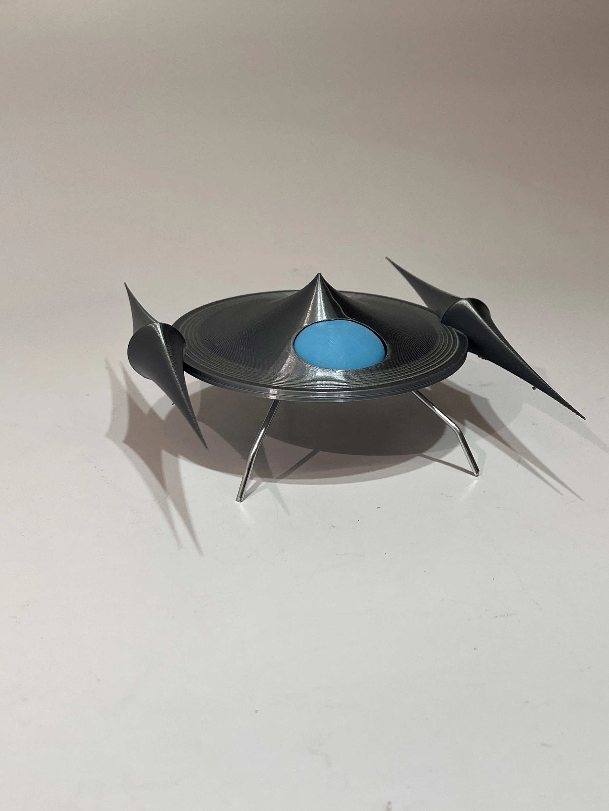 Small model spaceship with a black saucer-shaped body, three metal legs, and three sharp black fins on the sides.