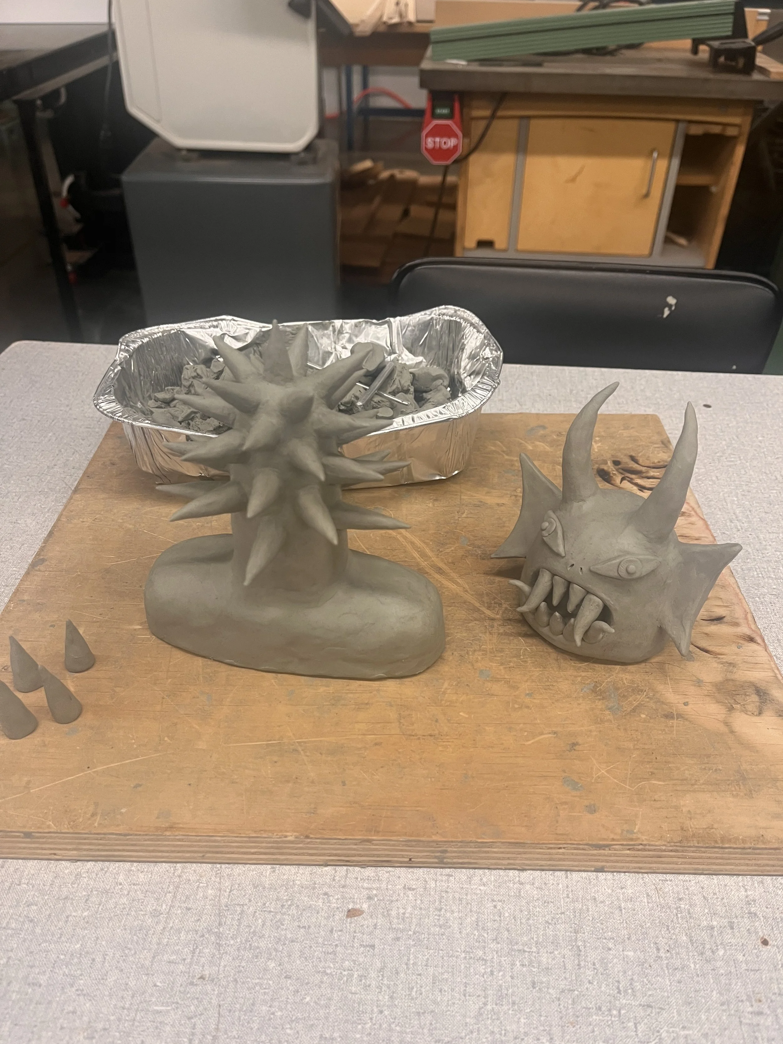 Two fantasy creature sculptures in progress, one with multiple spikes on its head and the other with horns and a menacing facial expression, placed on a wooden work surface. Clay tools and a foil tray are nearby.