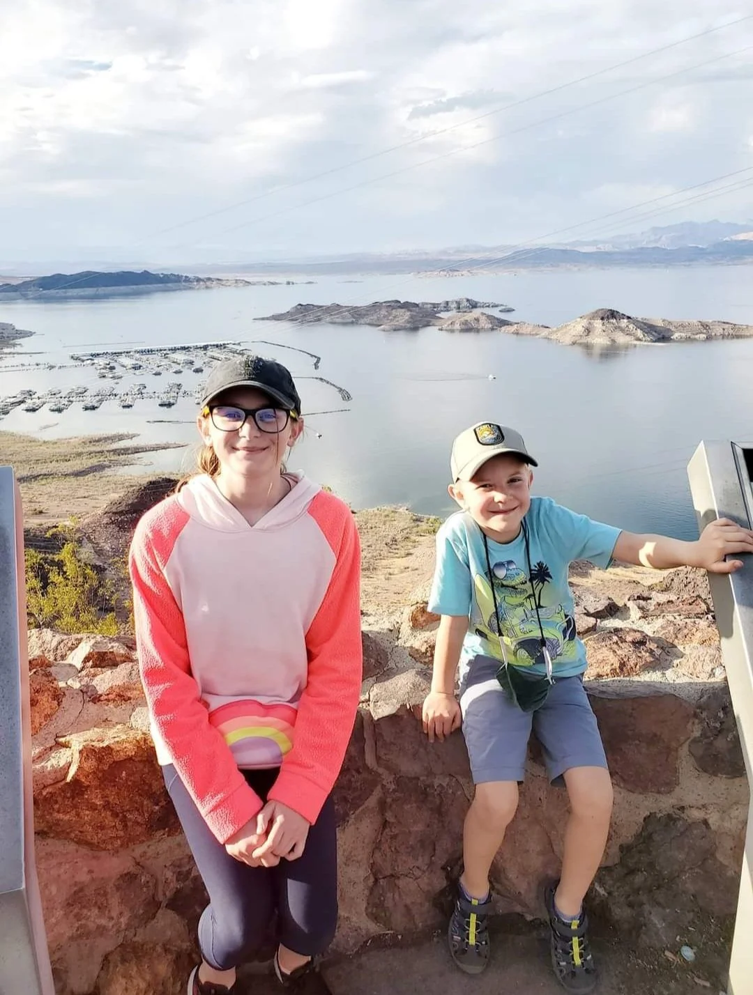 A girl and a boy standing outdoors, with a lake and hills in the background. The girl is smiling, wearing glasses, a black cap, with long reddish hair, pink and white hoodie, and gray leggings. The boy is smiling, wearing a baseball cap, a light blue T-shirt with a graphic, gray shorts, and sneakers. He is leaning on a rock formation, with a scenic view of water, islands, and mountains in the distance.