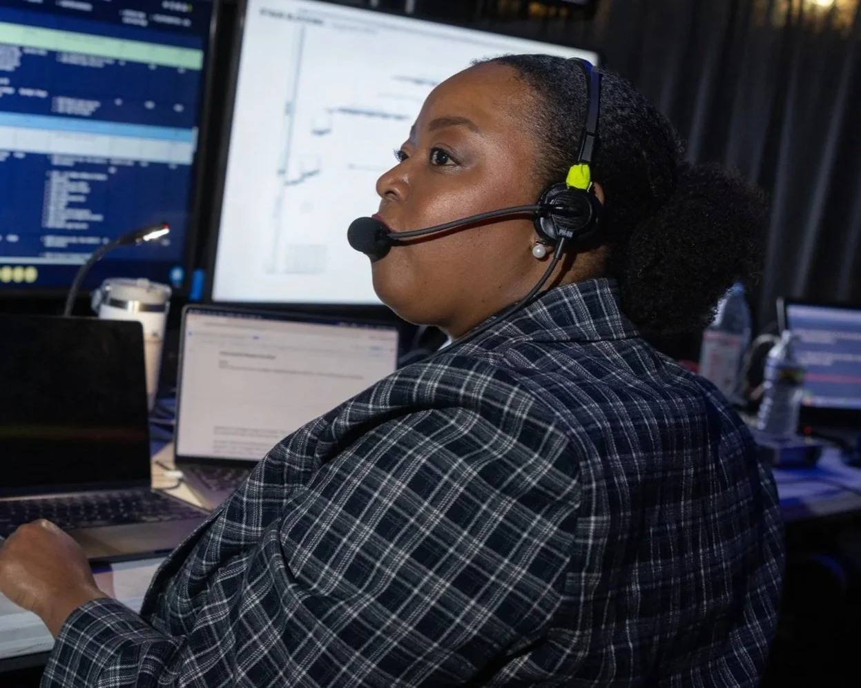 A woman wearing a headset with a microphone, sitting at a desk with multiple computer screens, working in an office or control room environment.