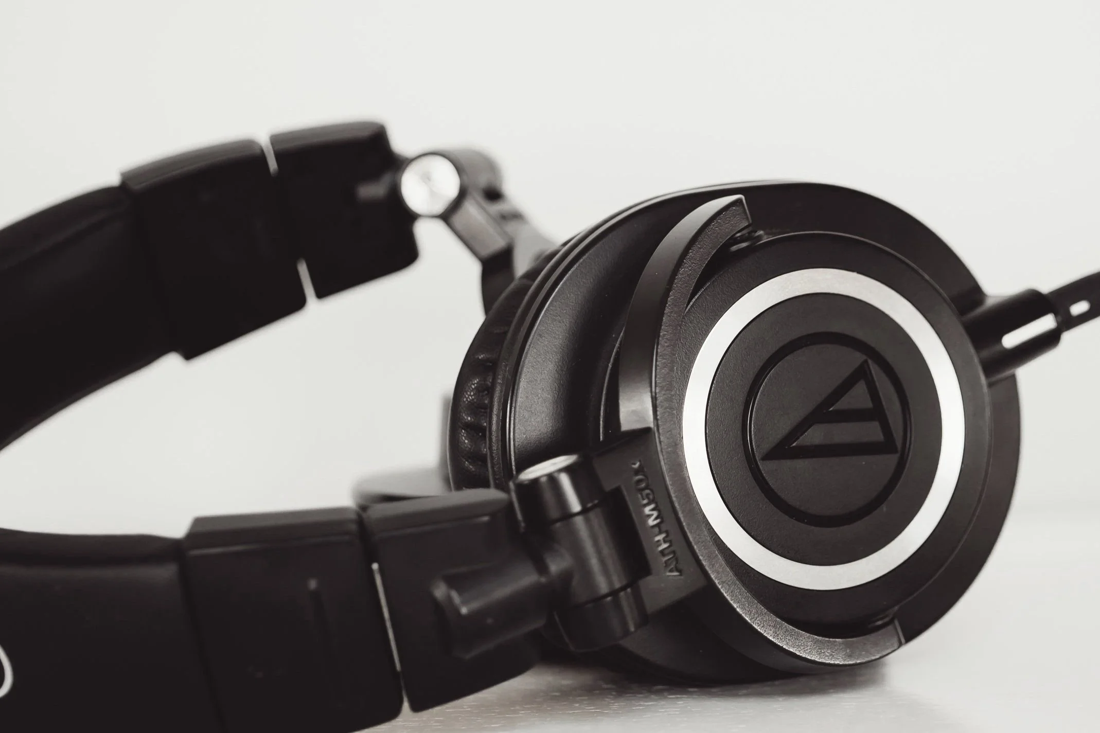 Close-up of a black over-ear headphone with a silver and black circular logo on the ear cup, set against a plain light background.