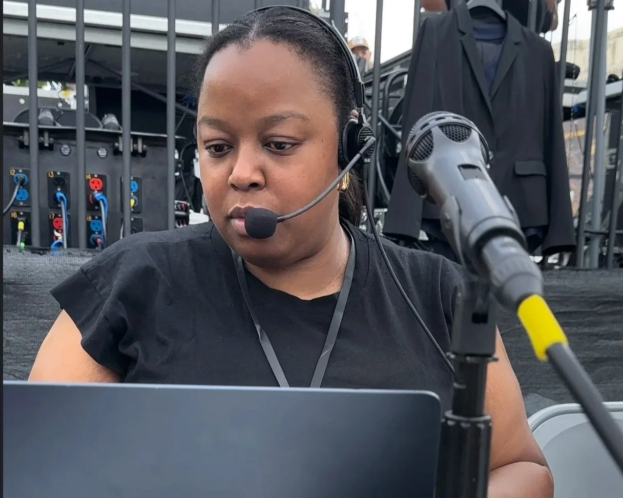 A woman wearing a headset with a microphone working on a laptop at an event, with audio equipment and hanging clothes in the background.