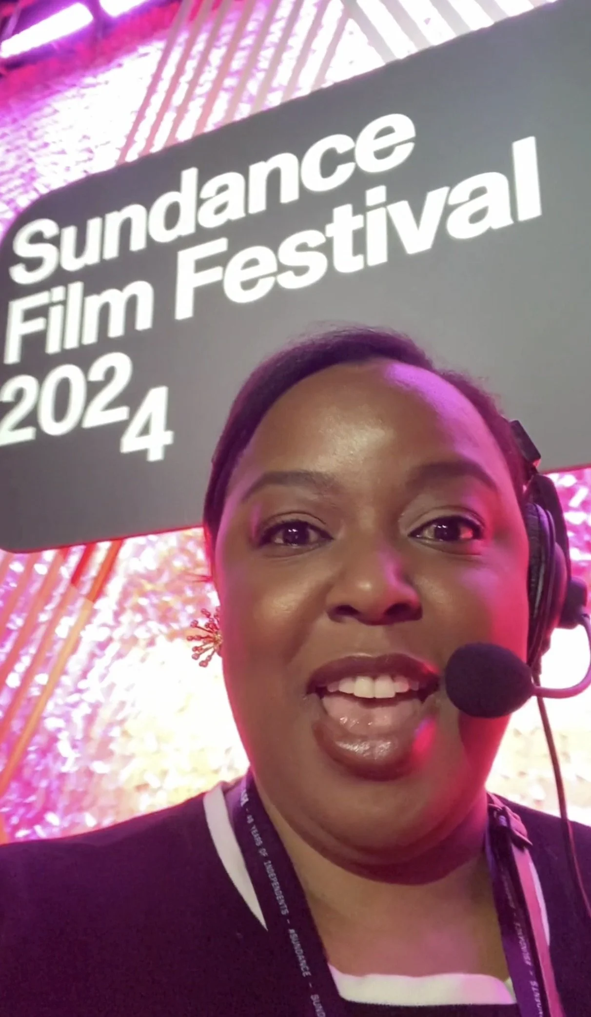 A woman smiling, wearing a headset with a microphone, in a setting with a sign that reads "Sundance Film Festival 2024."