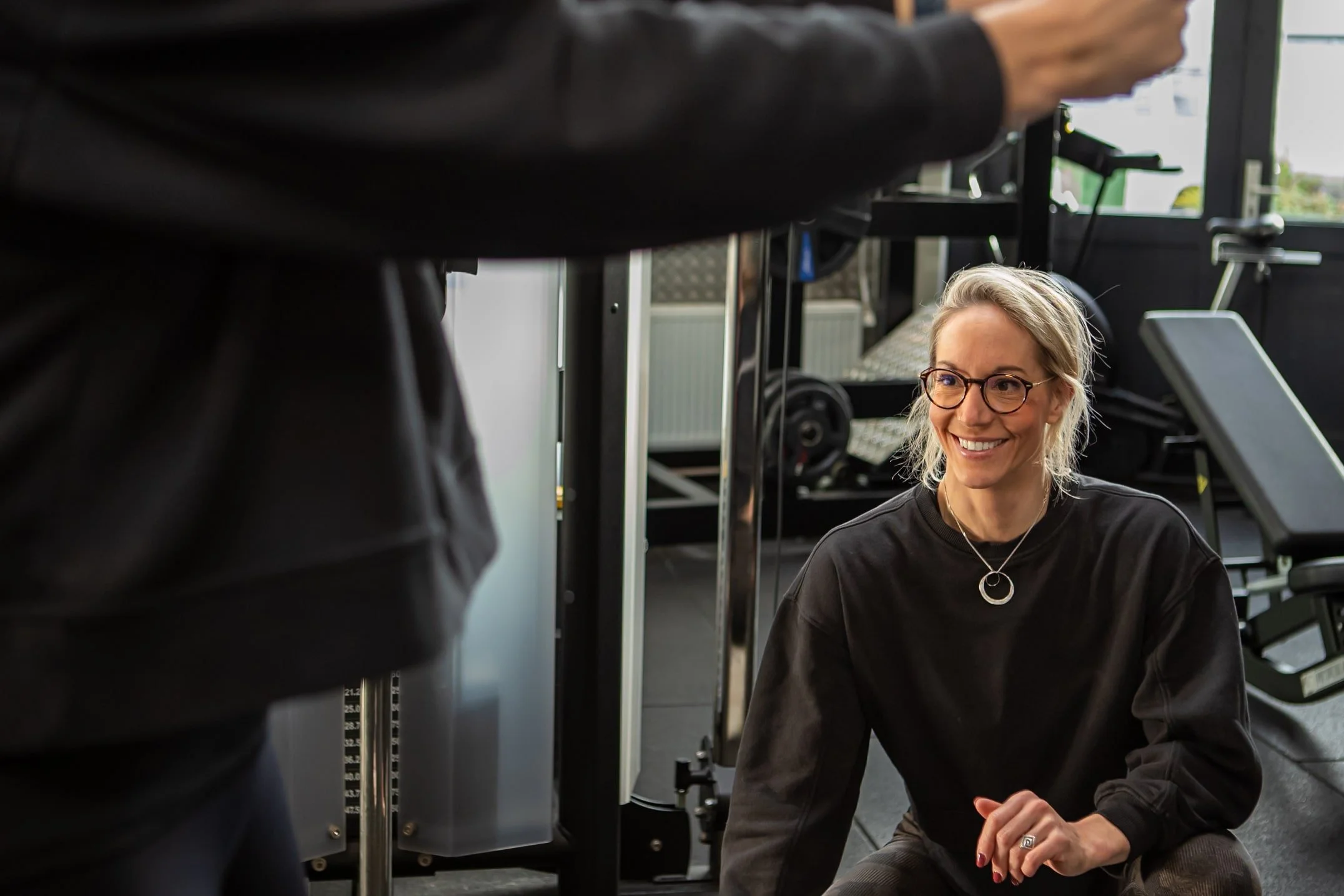 Woman with glasses in a gym setting, smiling, wearing a black sweatshirt, with gym equipment in the background.