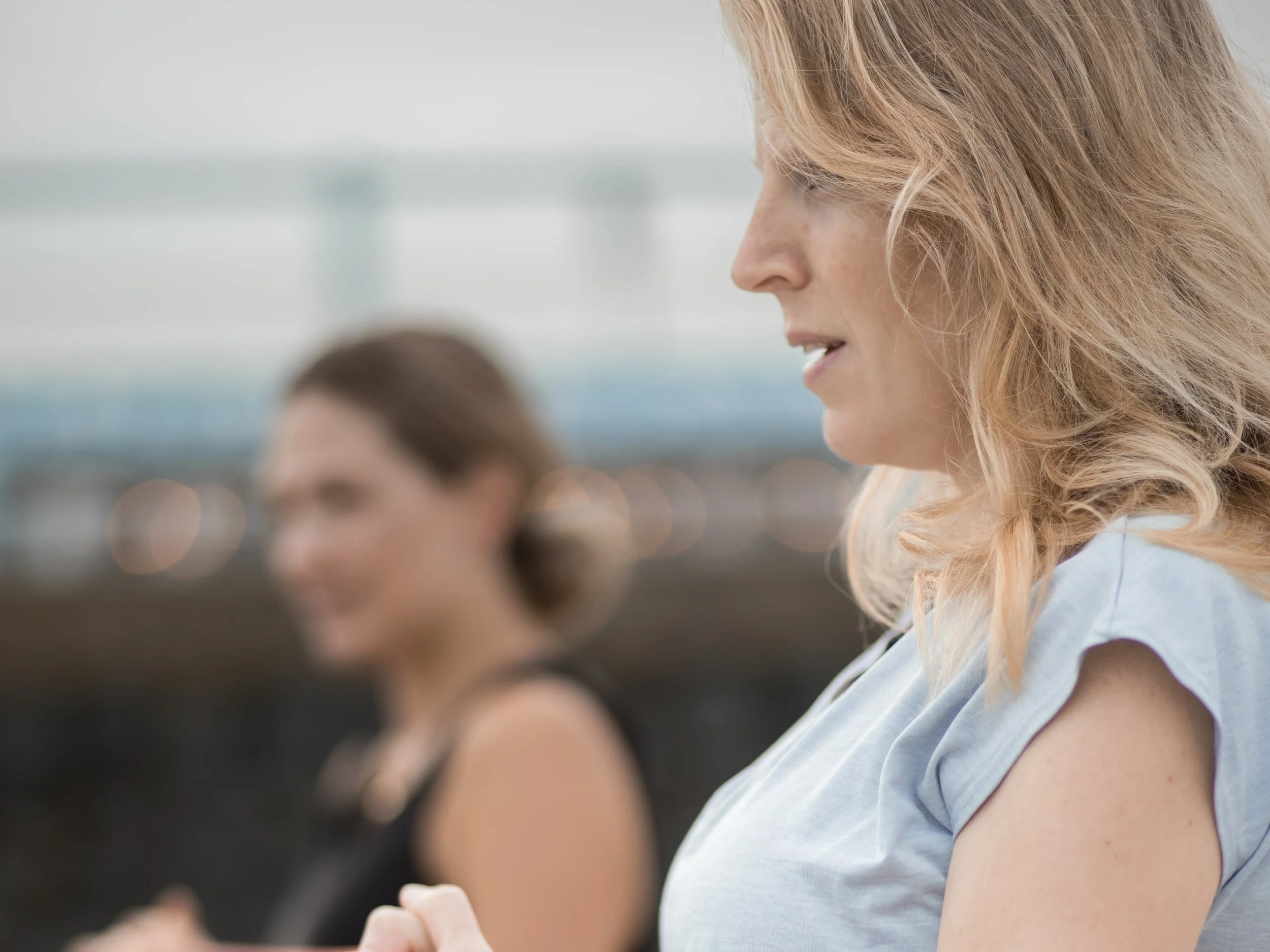 Two women standing outdoors, one in focus in the foreground and the other blurred in the background.