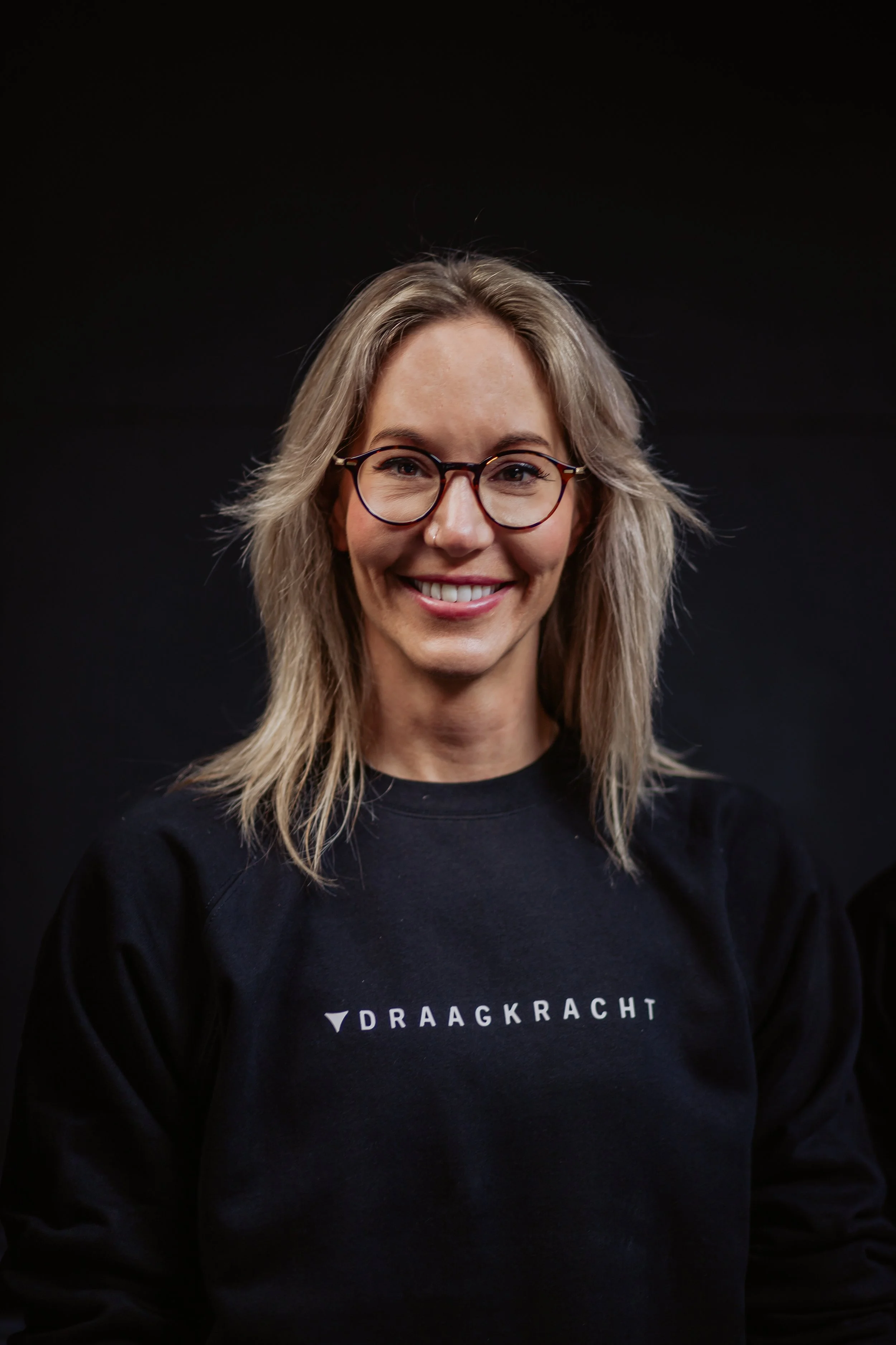 A smiling woman with glasses, blonde hair tied in a half-up ponytail, wearing a black workout tank top and leggings, holding dumbbells in a gym