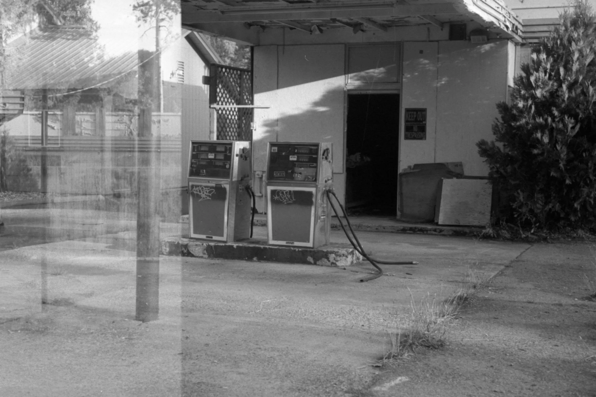 Black and white photo of a small building with a large open door and two gas pumps in front. There is a sign on the building that says 'STOP OUT'. Some trees and a fence are visible in the background.