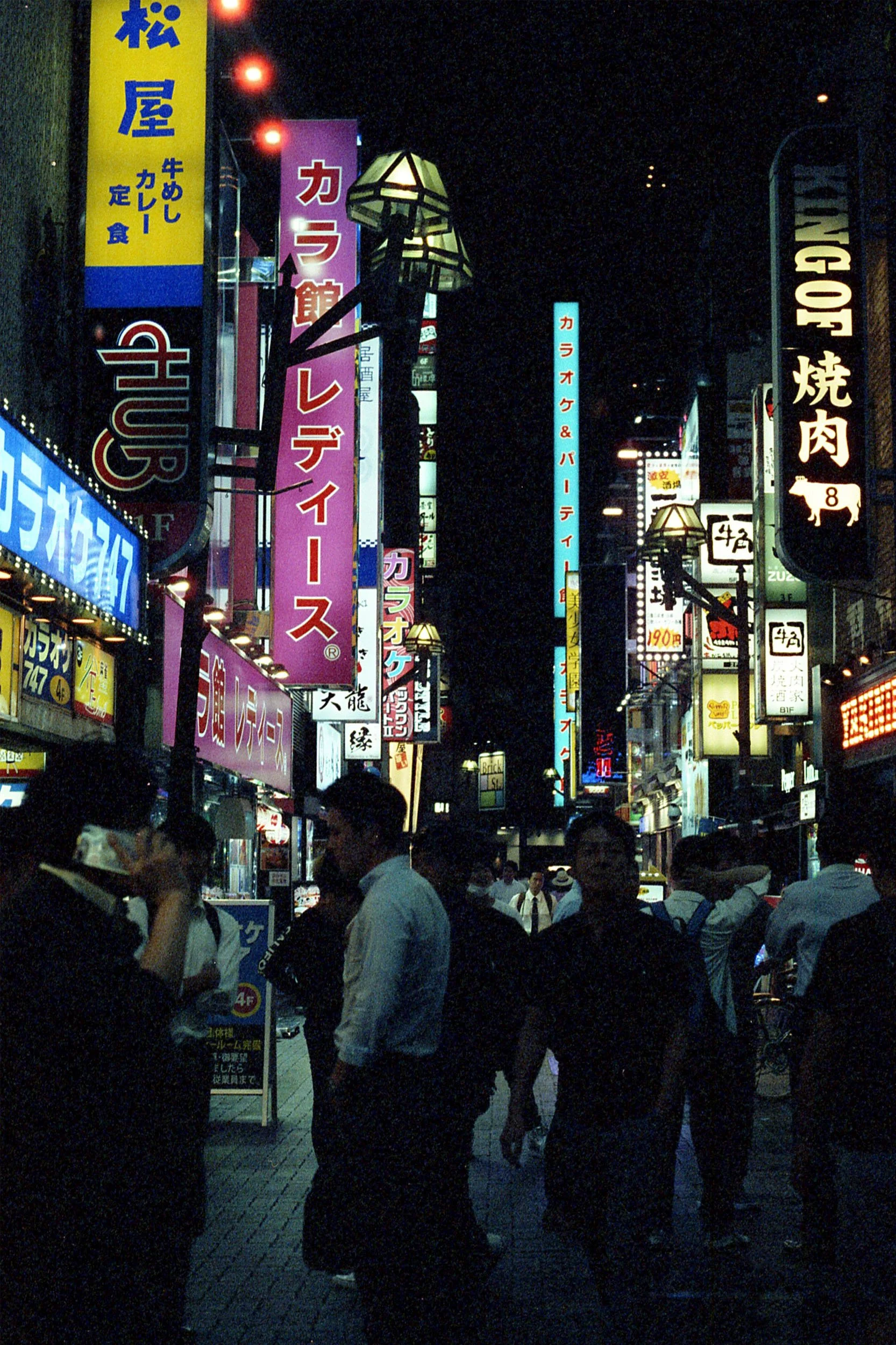 Nighttime scene of a busy street filled with illuminated signs in Japanese, people walking and standing around.
