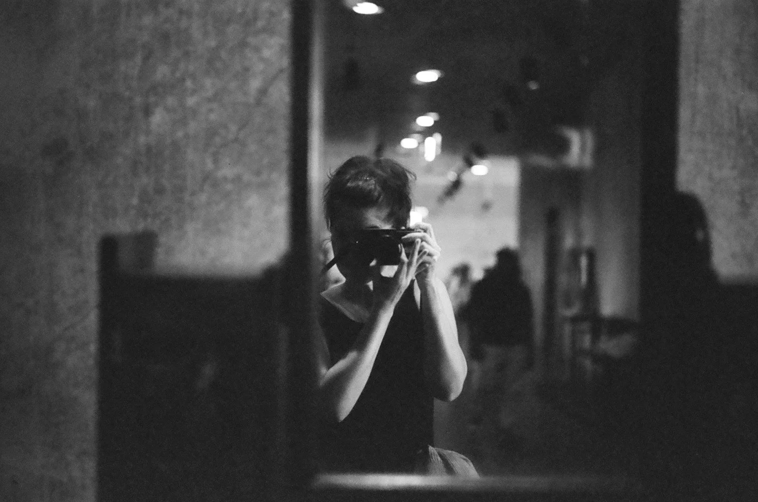 A person taking a photo in a mirror, holding a camera to their face, in a dimly lit, industrial-style hallway, black and white photograph