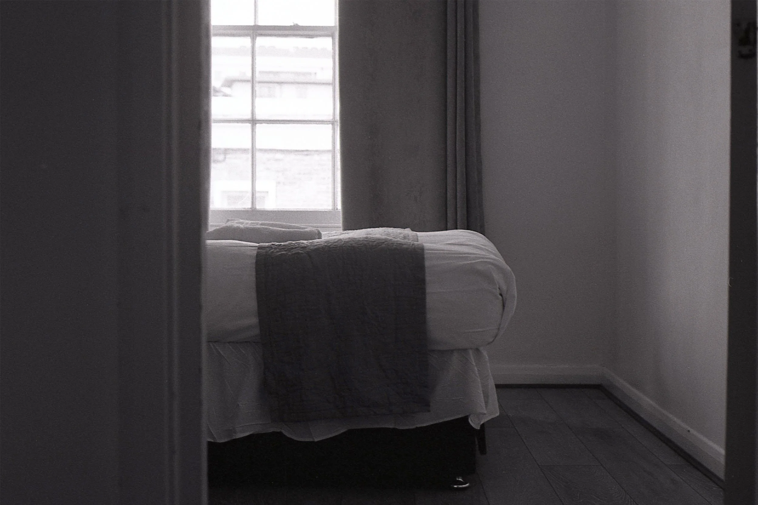 A bed with white bedding and a gray blanket in front of a window with white frames. The photo is taken through a doorway or mirror, creating a partial view of the room.
