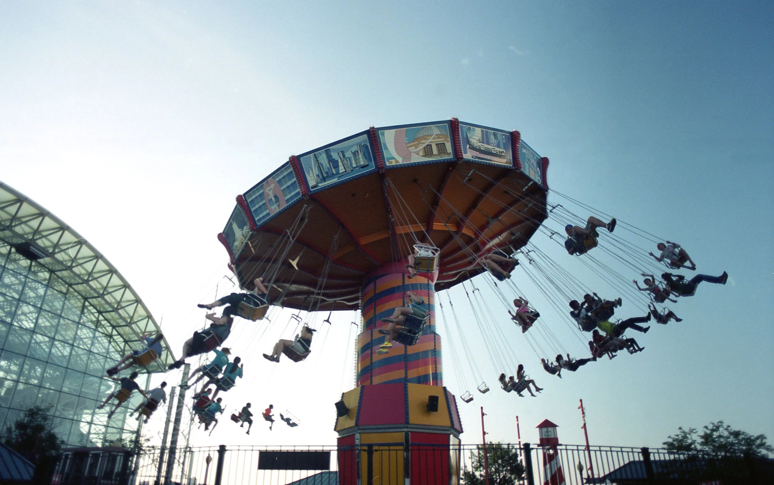 A colorful swing ride at Navy Pier, Chicago with children and adults riding, set against a clear blue sky with a large modern glass building in the background.