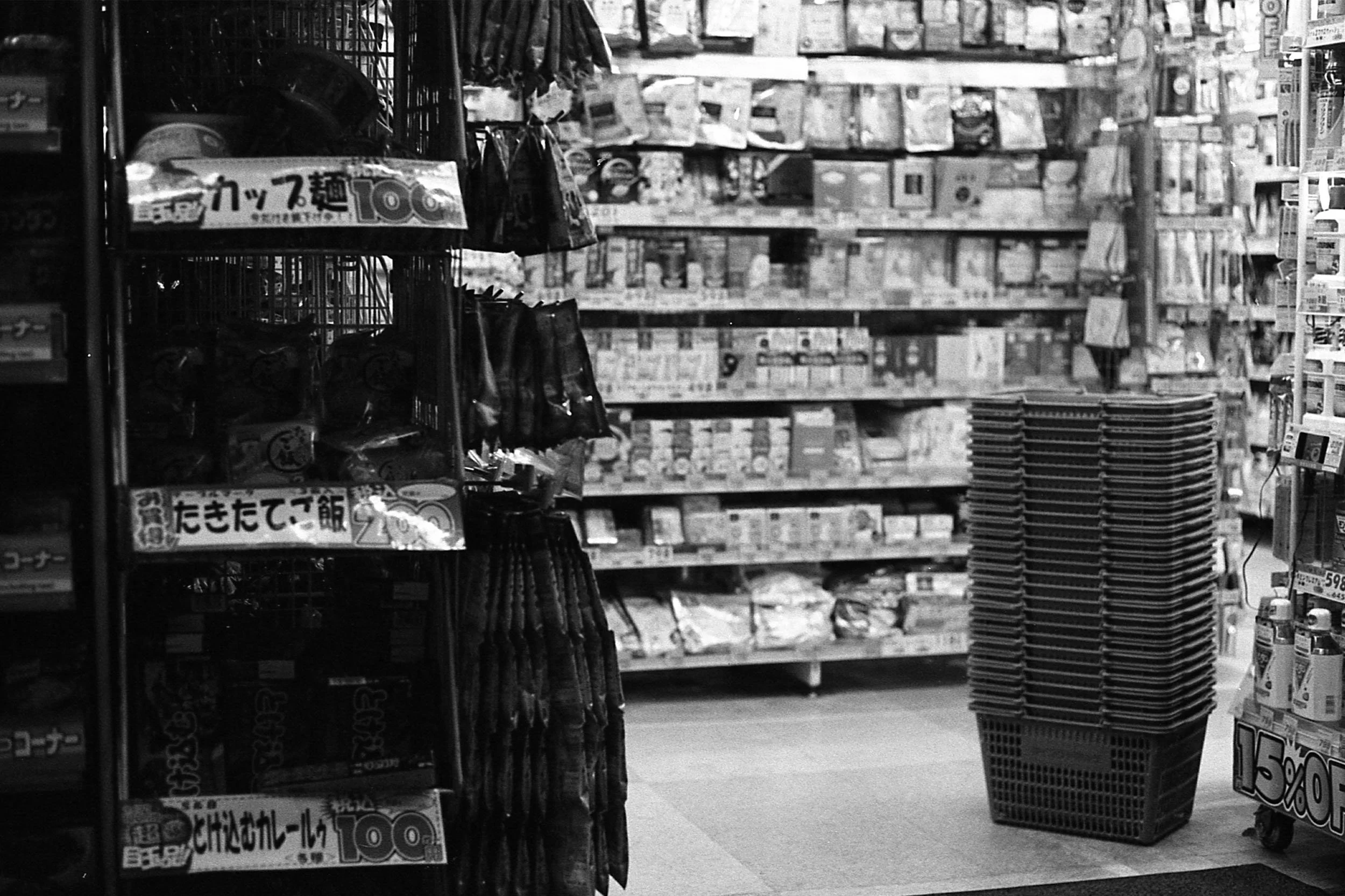 A black-and-white photo of an aisle in a grocery store with shelves stocked with various products. There are baskets full of items and some signage with Japanese text and prices.