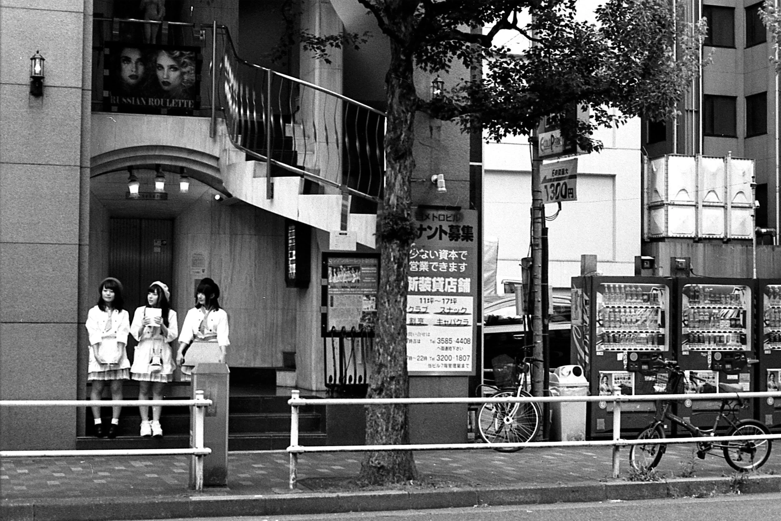 Three young women in matching white outfits standing on a sidewalk, with a tree, bicycle, advertising signs, and vending machines nearby in an urban setting.