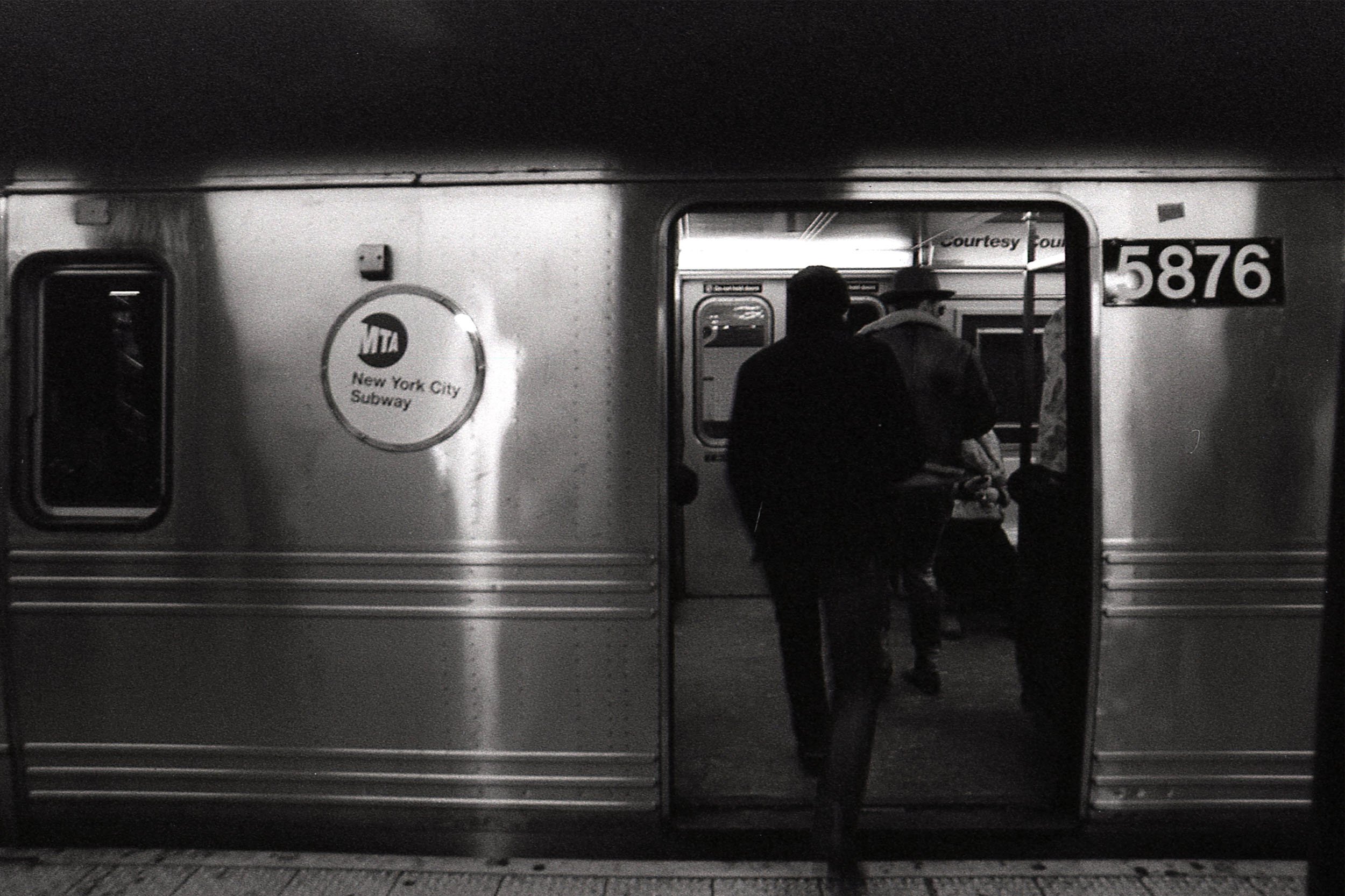 Black and white photo of two people boarding a New York City subway train through an open door, with visible subway car number 5876.