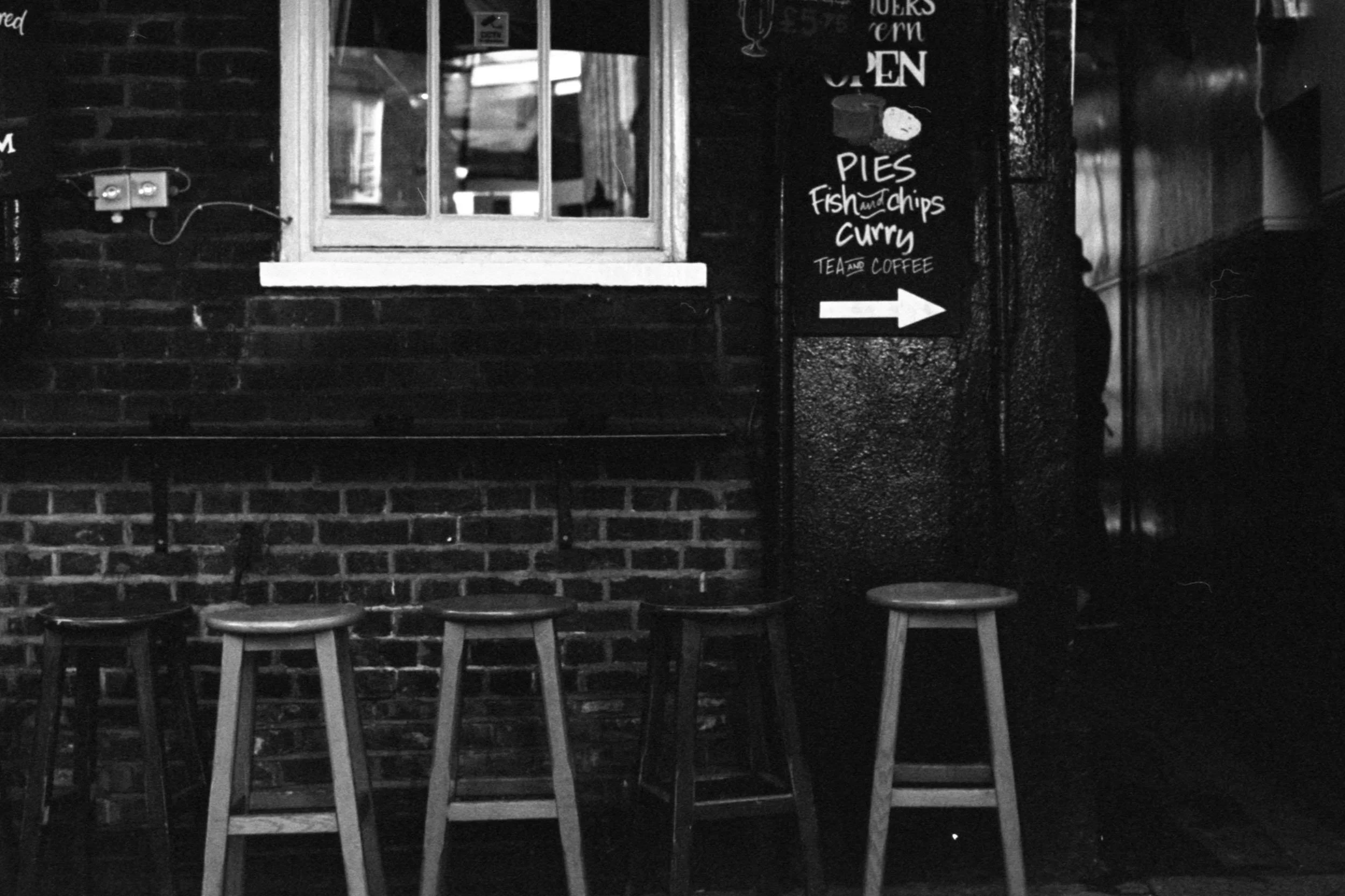 Black and white photo of a cafe with a brick wall, stools, a window, and a chalkboard sign mentioning pies, fish and chips, curry, tea and coffee, with an arrow pointing to the right.