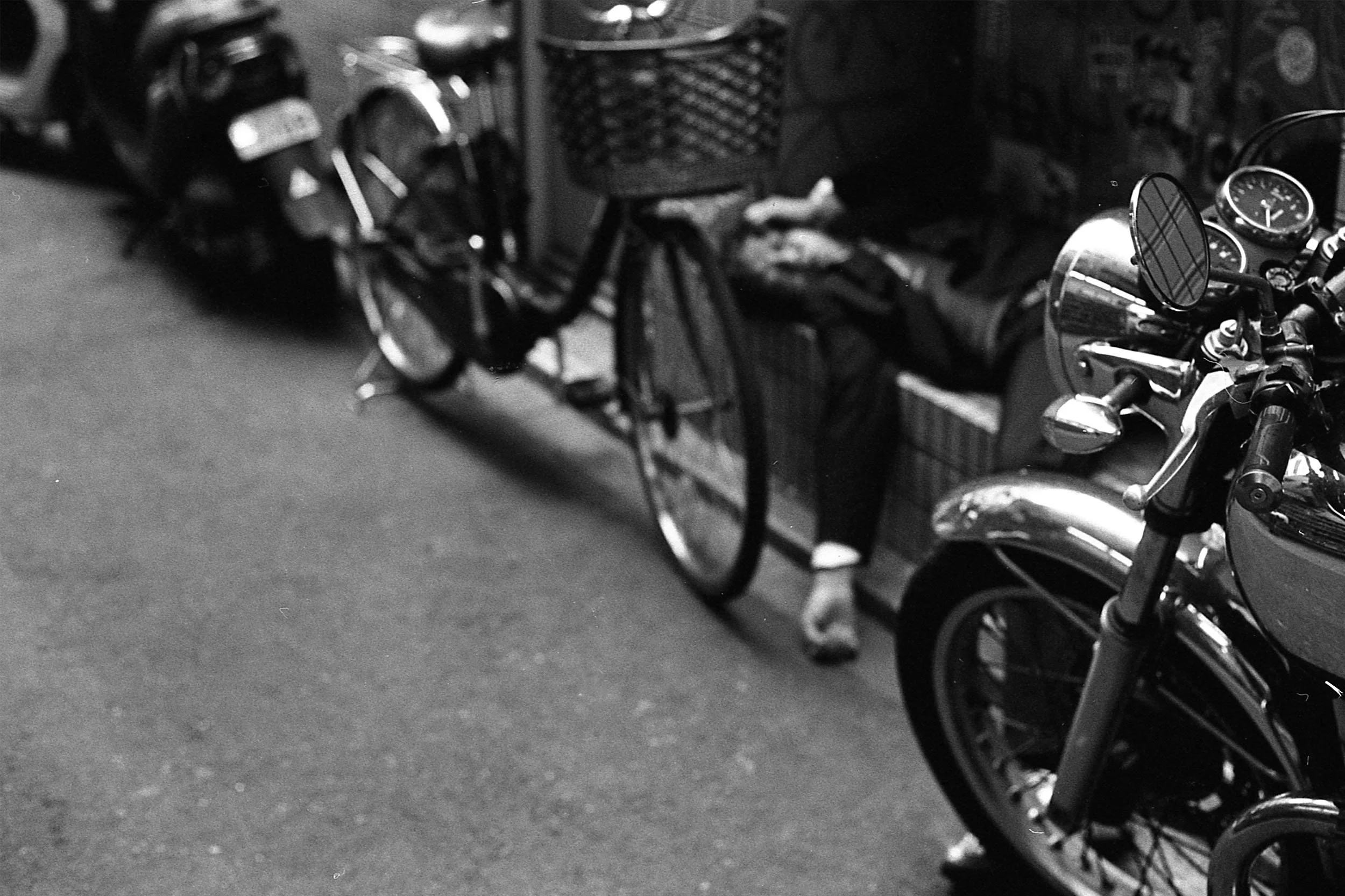 Black and white photograph of a motorcycle parked indoors, with its front part and handlebars visible, a man in a suit is passed out on the sidewalk behind.
