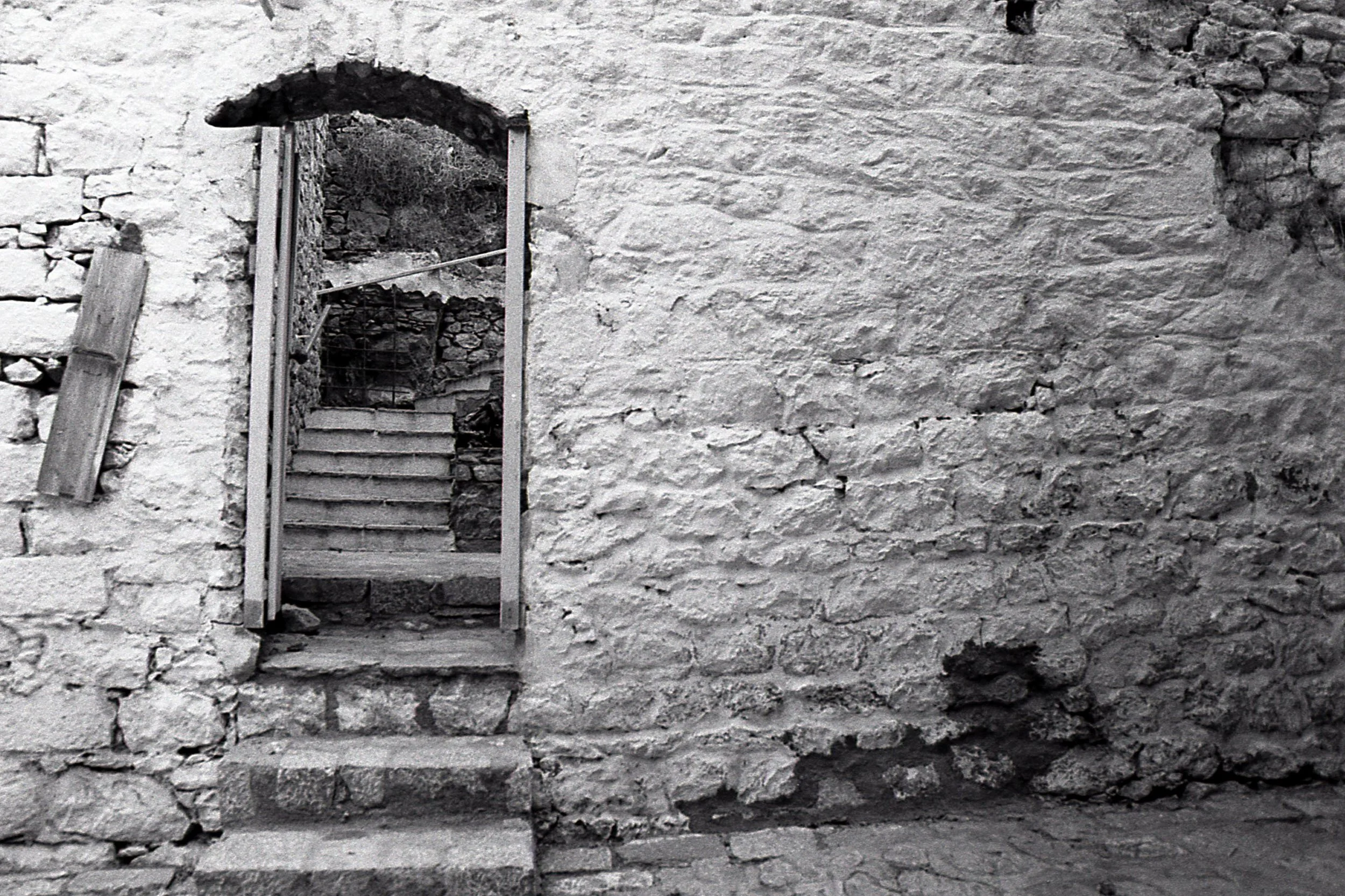 Old stone wall with a small opening showing stairs and plants through it.