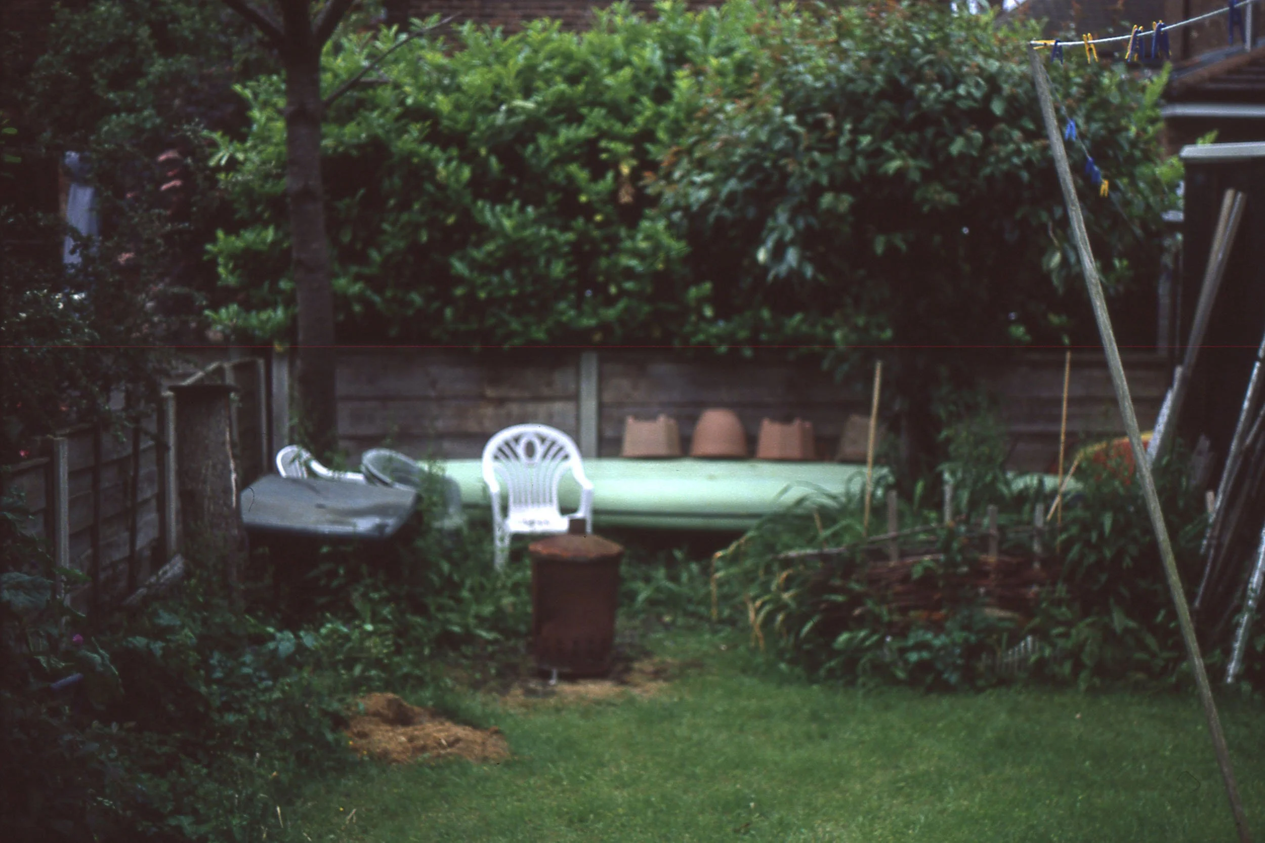 A backyard garden with a table, white plastic chairs, potted plants, and a clothesline with clothespins. There are trees and bushes surrounding the area, with a wooden fence in the background.
