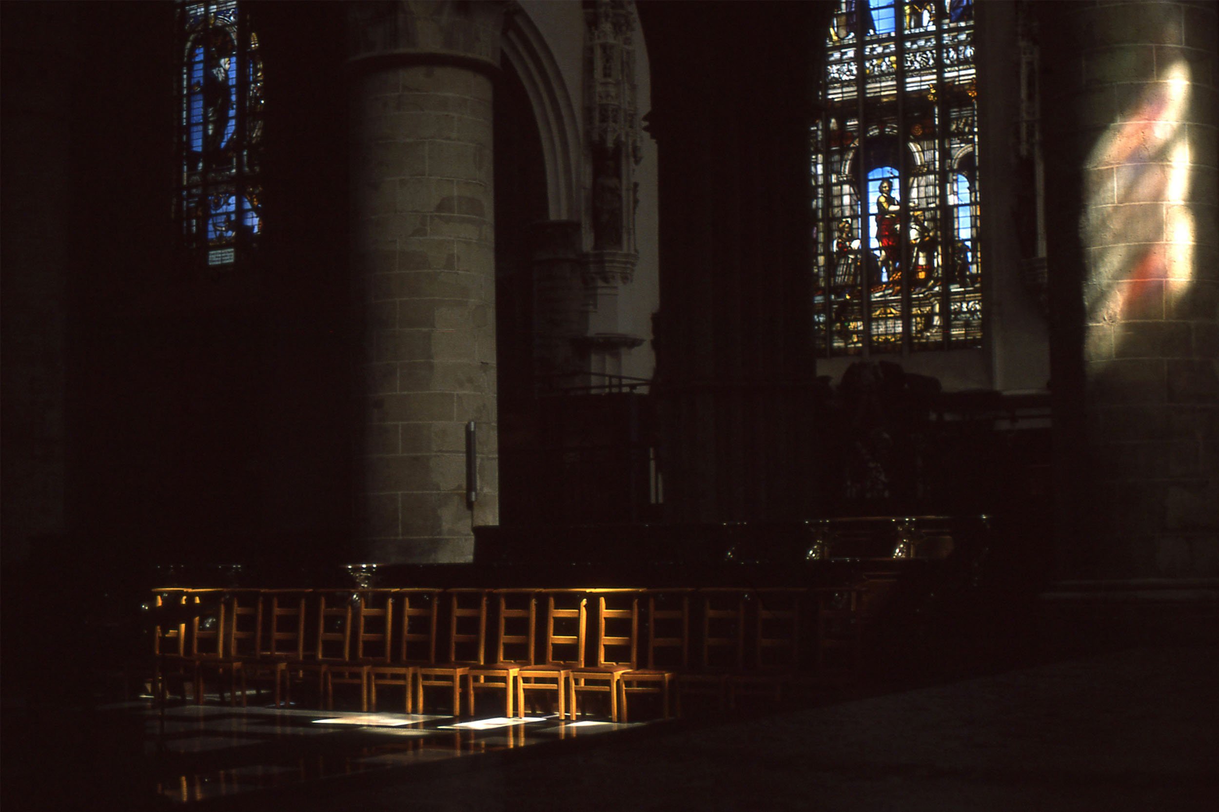 Interior of a church with stained glass windows, stone columns, and a lit altar with a row of empty chairs.