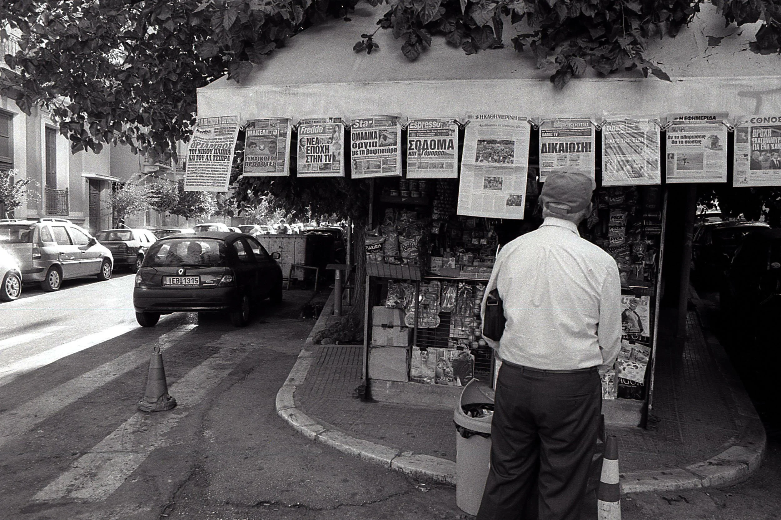 A man in a white shirt and dark pants stands near a newsstand on a city sidewalk, with cars parked along the street behind him. The newsstand sells newspapers, magazines, and snacks, with several newspapers hanging on display.