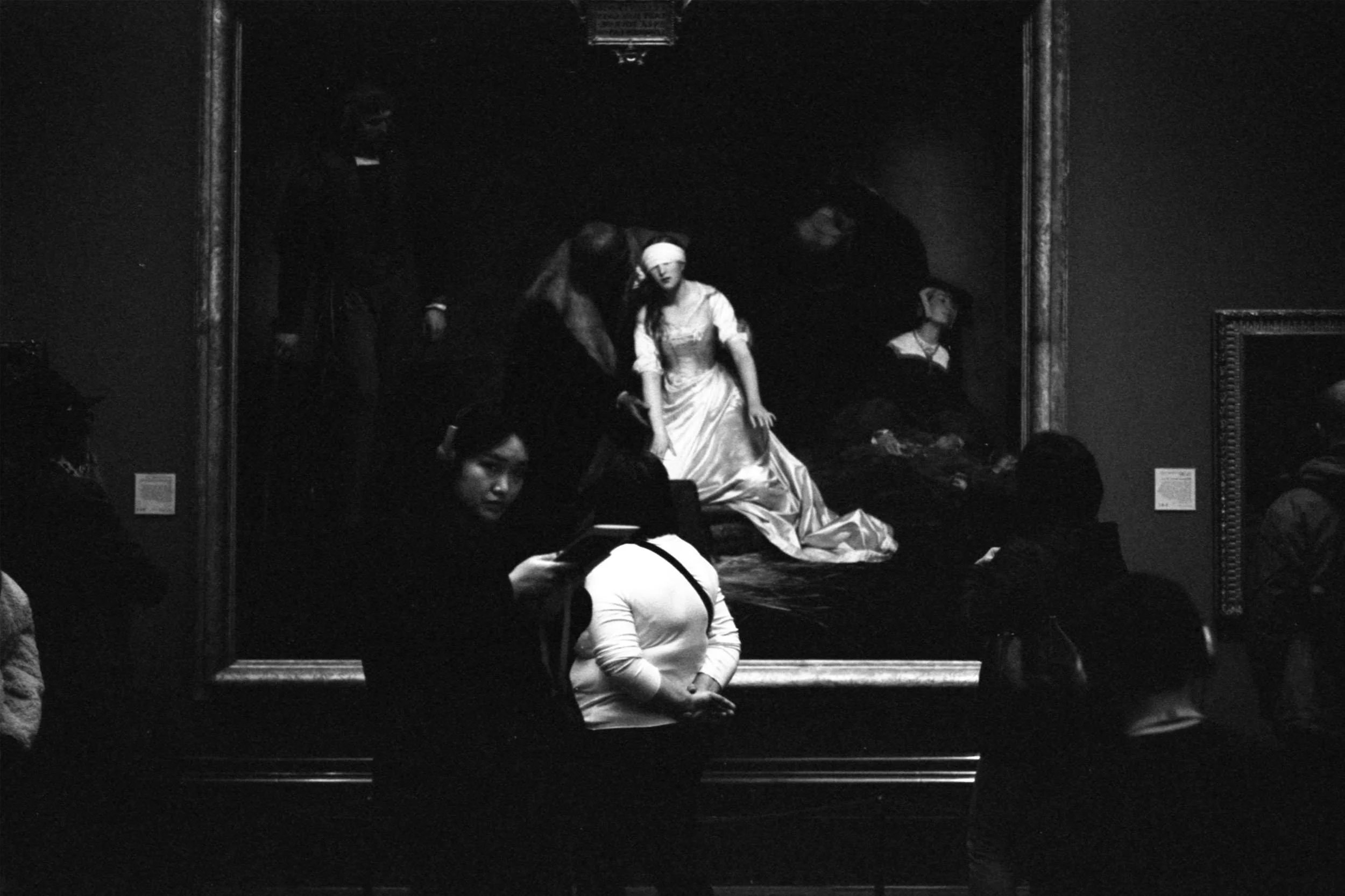 Black and white photo of a painting hanging in a gallery with people sitting in front of it, a woman with a bandage on her head in a fancy dress at the center, and visitors observing and engaging with the artwork.
