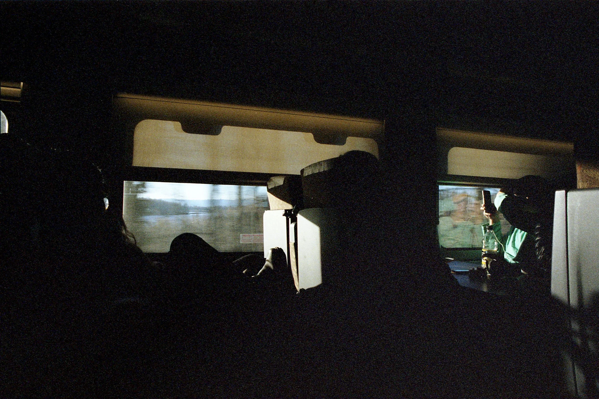 The image shows the interior of a vehicle at sunset, with silhouettes of three people — two on the left and one on the right. The person on the right is wearing a green shirt and is holding a smartphone, possibly taking a photo or video. The background visible through the window includes a roadside and some trees.