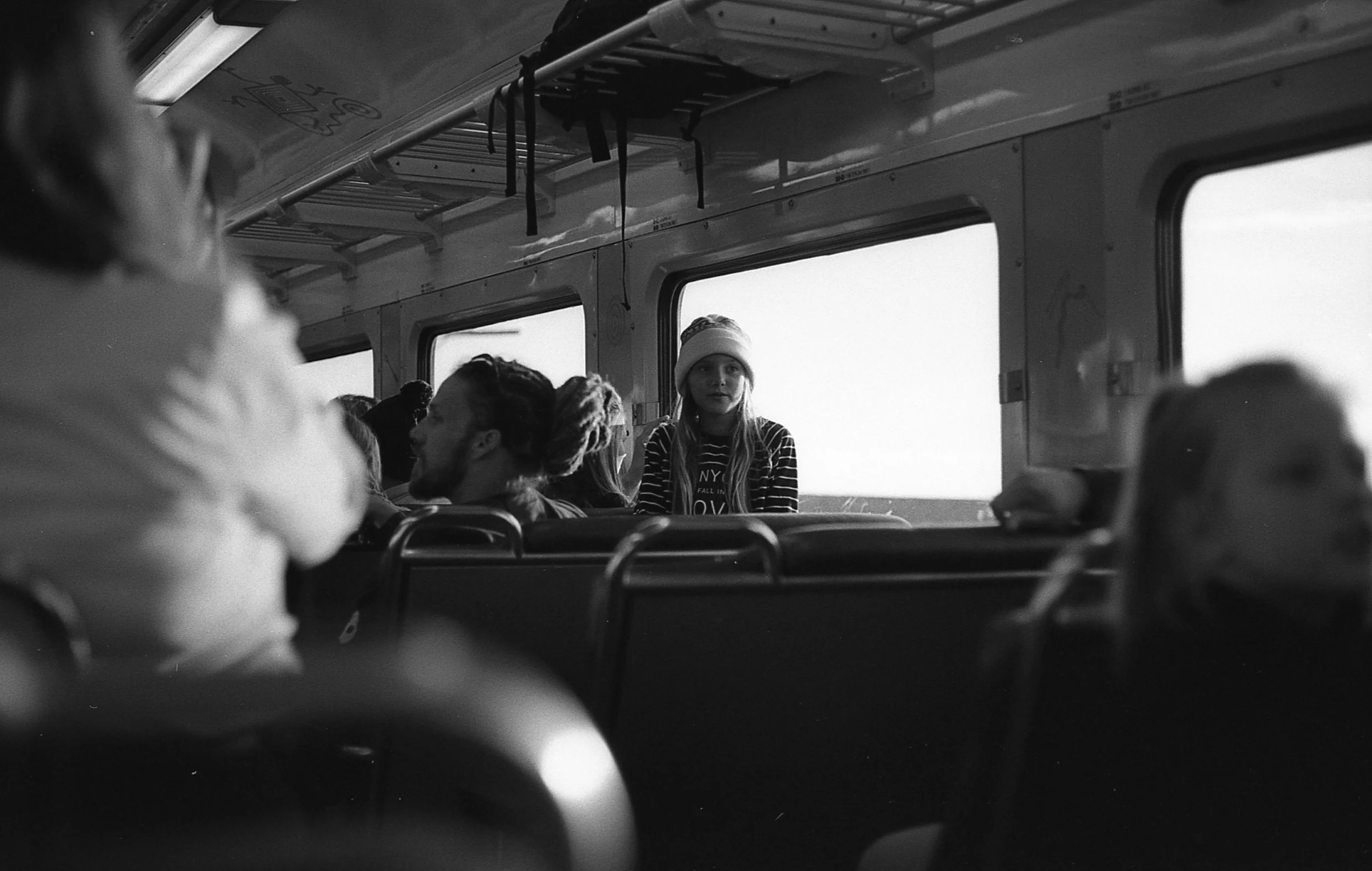 Black and white photo of the interior of a bus with several passengers seated, a young woman with a beanie sitting near the window, and other passengers engaging in conversations or looking forward.