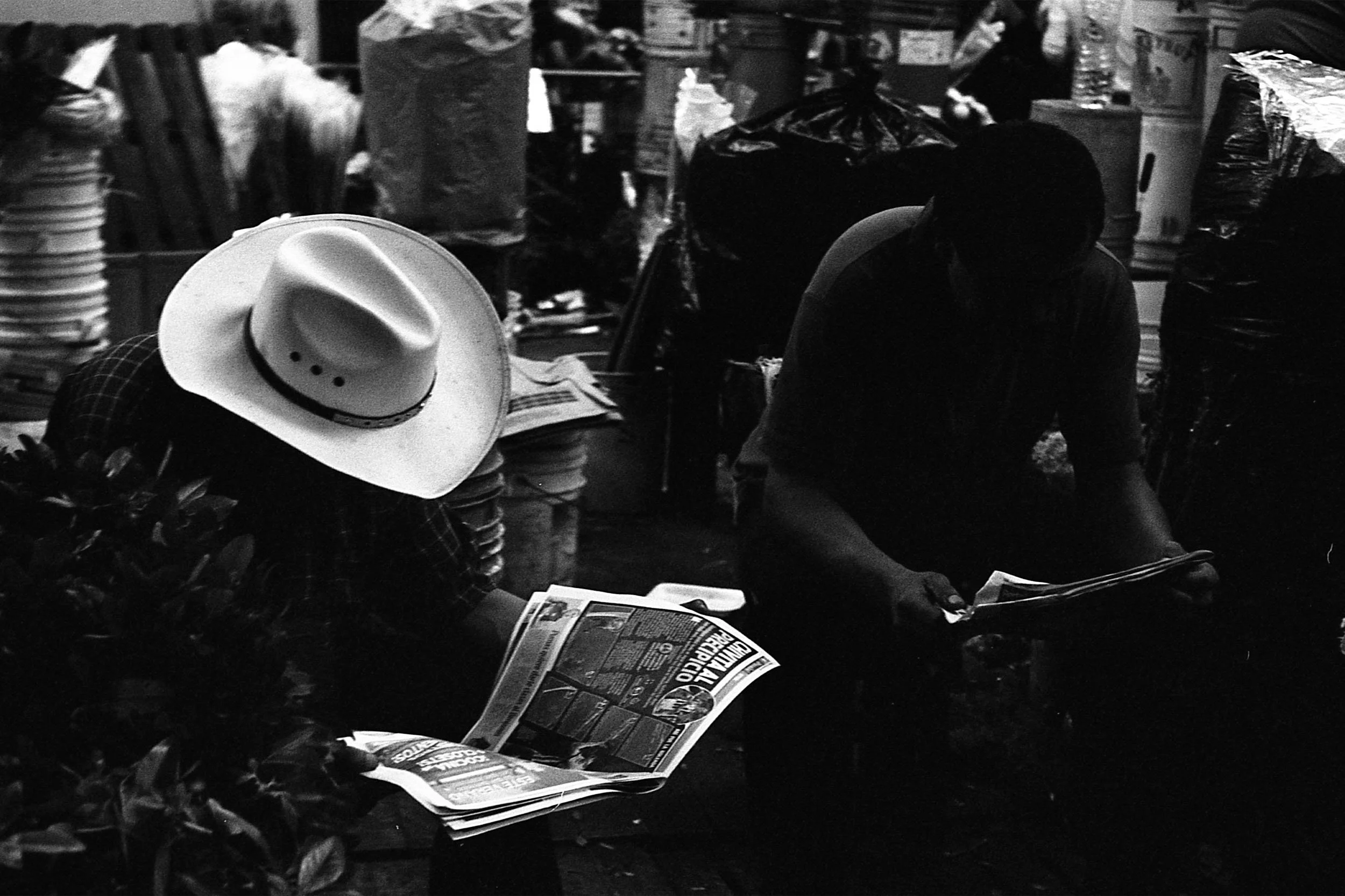 Black and white photo of two men reading newspapers. One in a large white cowboy hat.