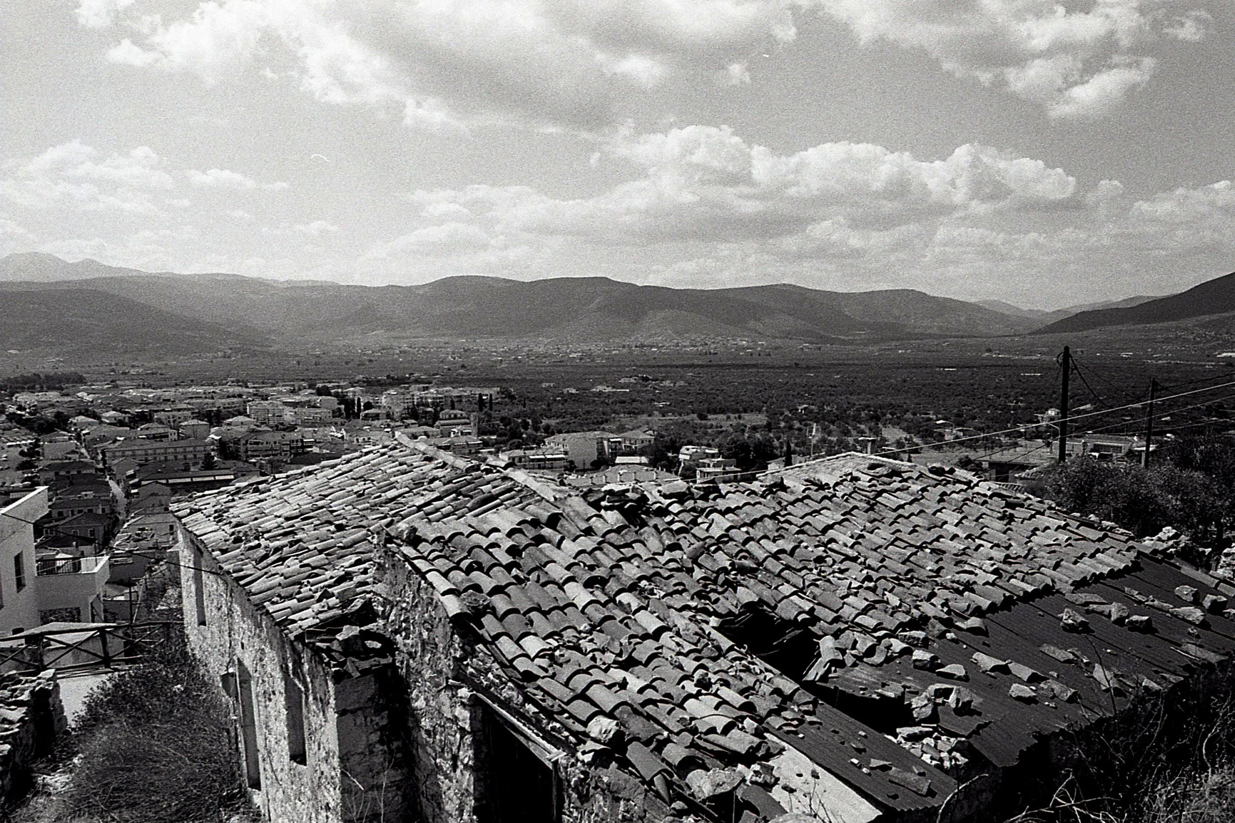 A black and white photo of a town with tiled roofs in the foreground, behind which are hills and mountains under a cloudy sky.