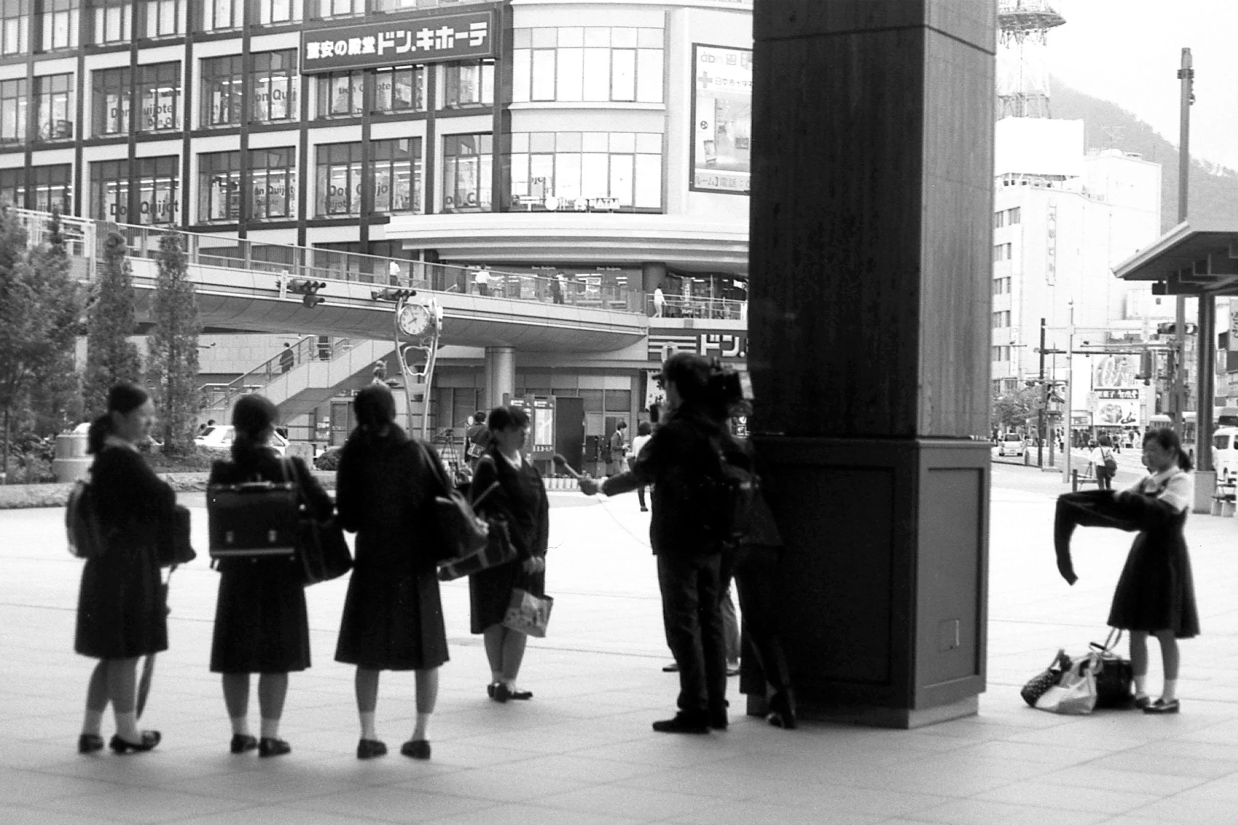 Black and white photo of a group of women dressed in school uniforms waiting in line at a train station in Japan. They are carrying bags, with some engaging in conversation. The station has a large building in the background with multiple floors and signs in Japanese, including a Don Quijote store. There is a clock and various trains or buses visible in the background.