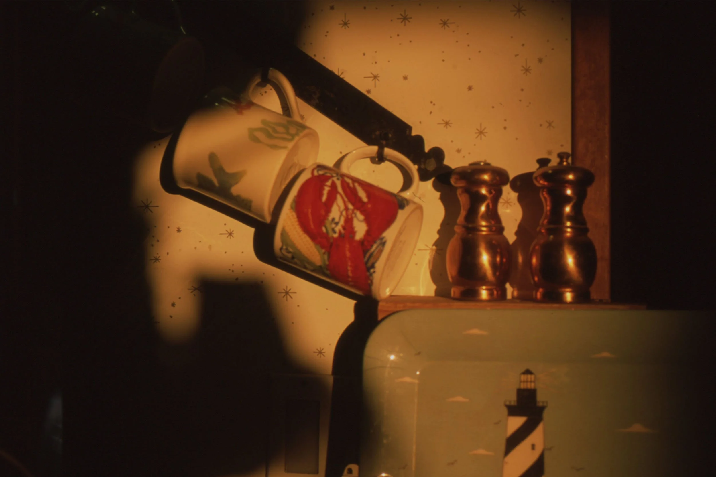 A kitchen shelf with two decorative mugs, a black dish brush, and three copper salt and pepper shakers, against a starry night sky wallpaper with lighthouse silhouette and clouds.