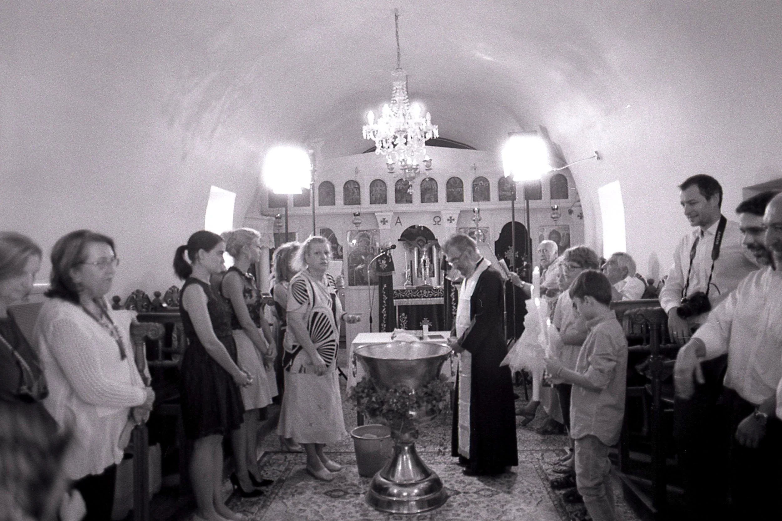 People participating in a religious ceremony inside a church, standing in two rows facing each other, with a priest or officiant in the center holding a book, and a large chandelier hanging from the ceiling.