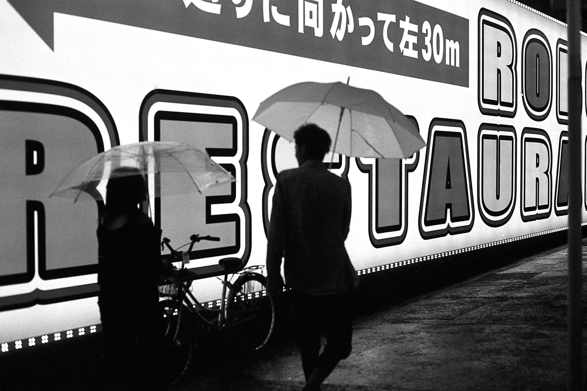 Two people holding umbrellas and standing next to bicycles in front of a large sign with Japanese writing and the word 'BICYCLE' in English.