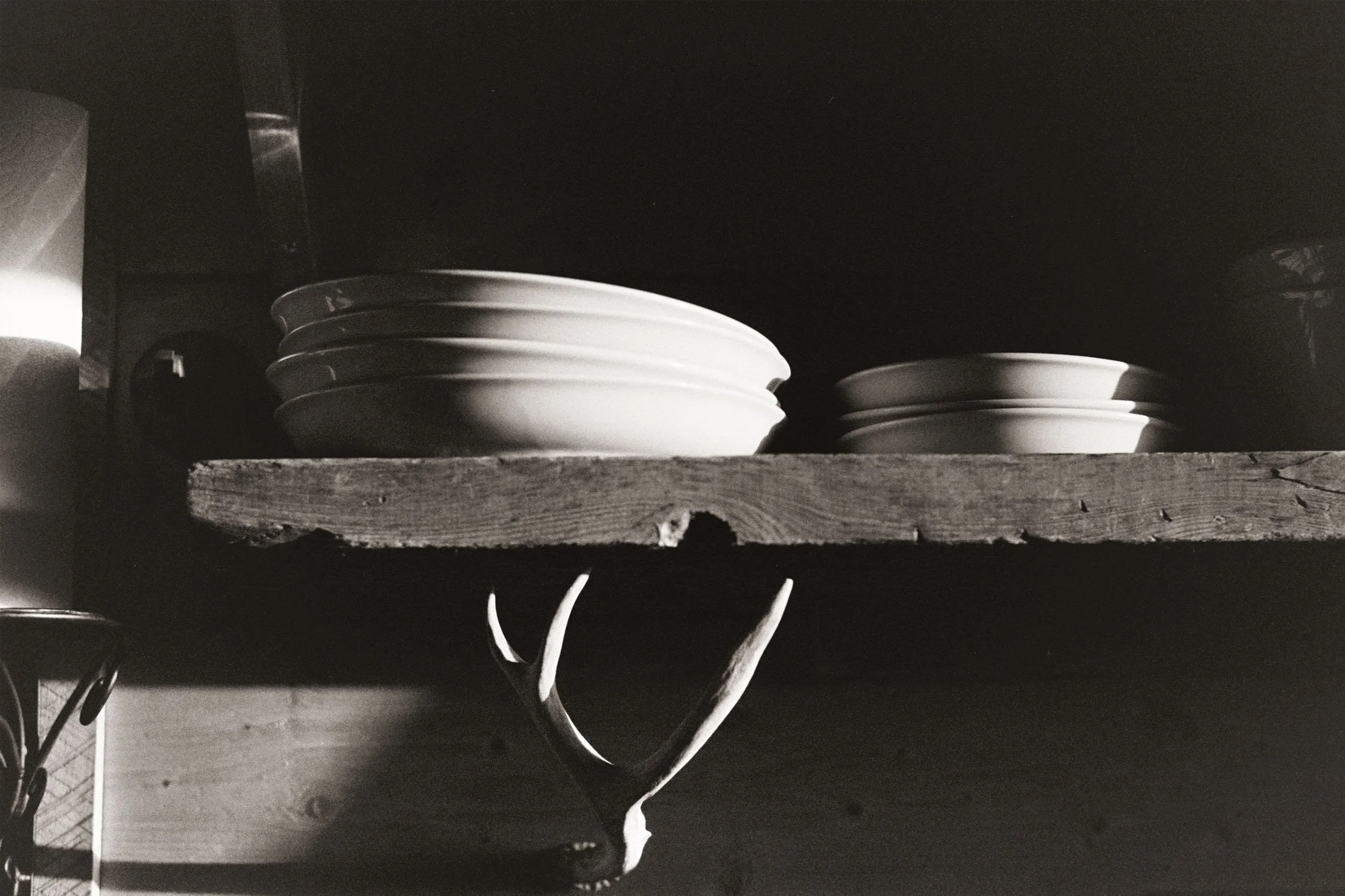 Black and white photo of a wooden shelf with several stacked white plates and some electrical cords underneath.