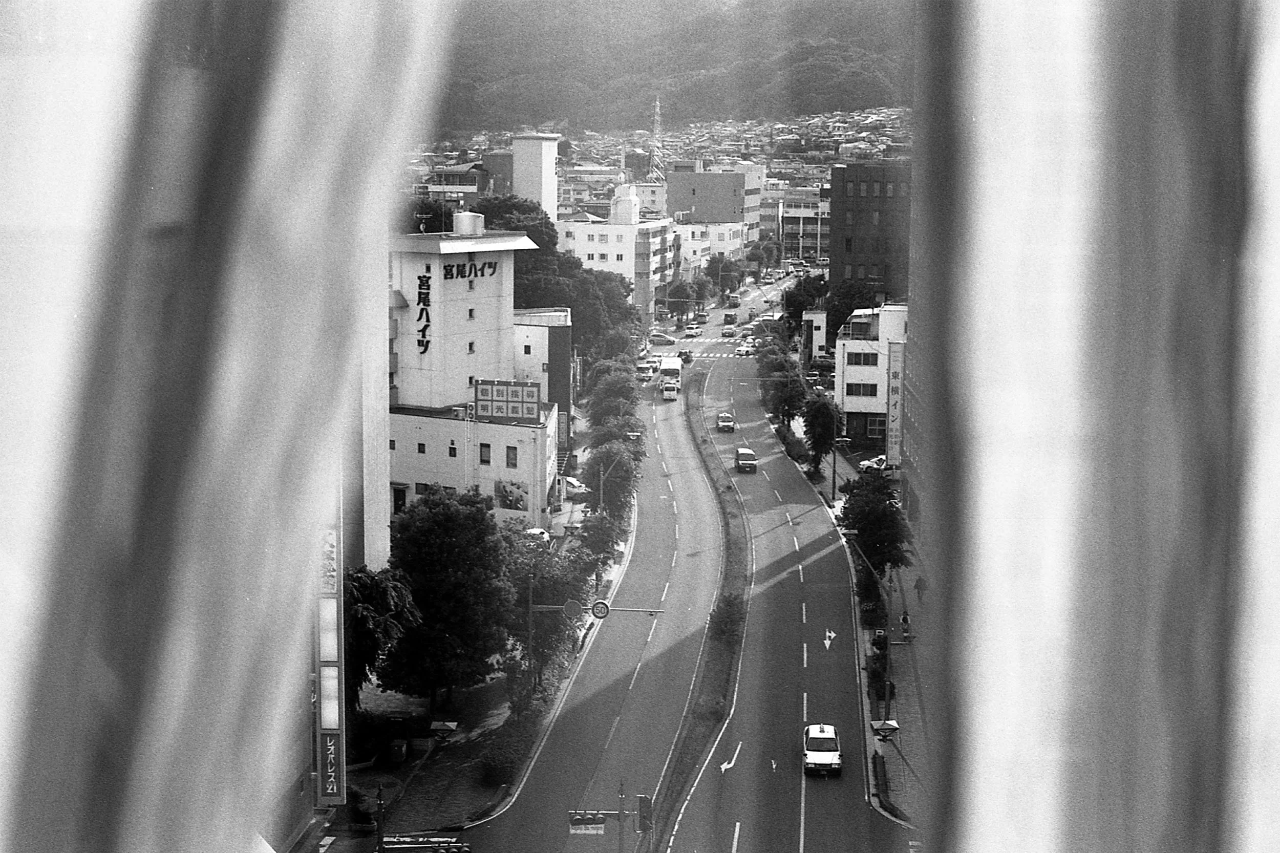 A black and white photo of a city street viewed between two vertical window blinds. The street is busy with cars, lined with trees and buildings, with a hilly background.