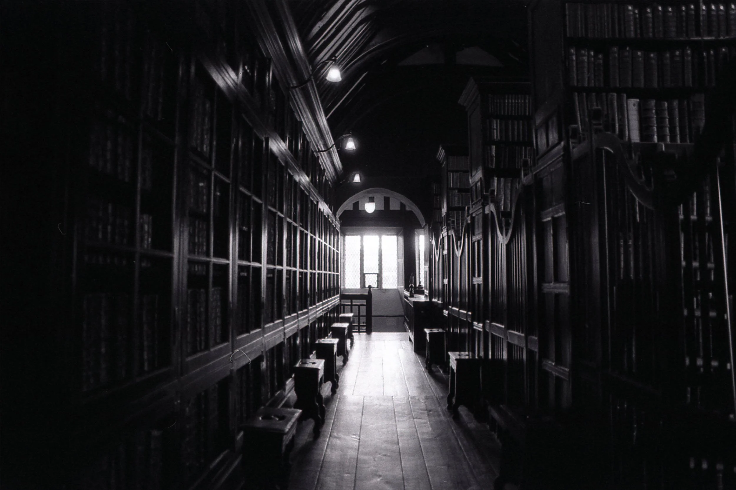 Dimly lit library with tall bookshelves on both sides, arched ceiling, and a window at the end letting in light.