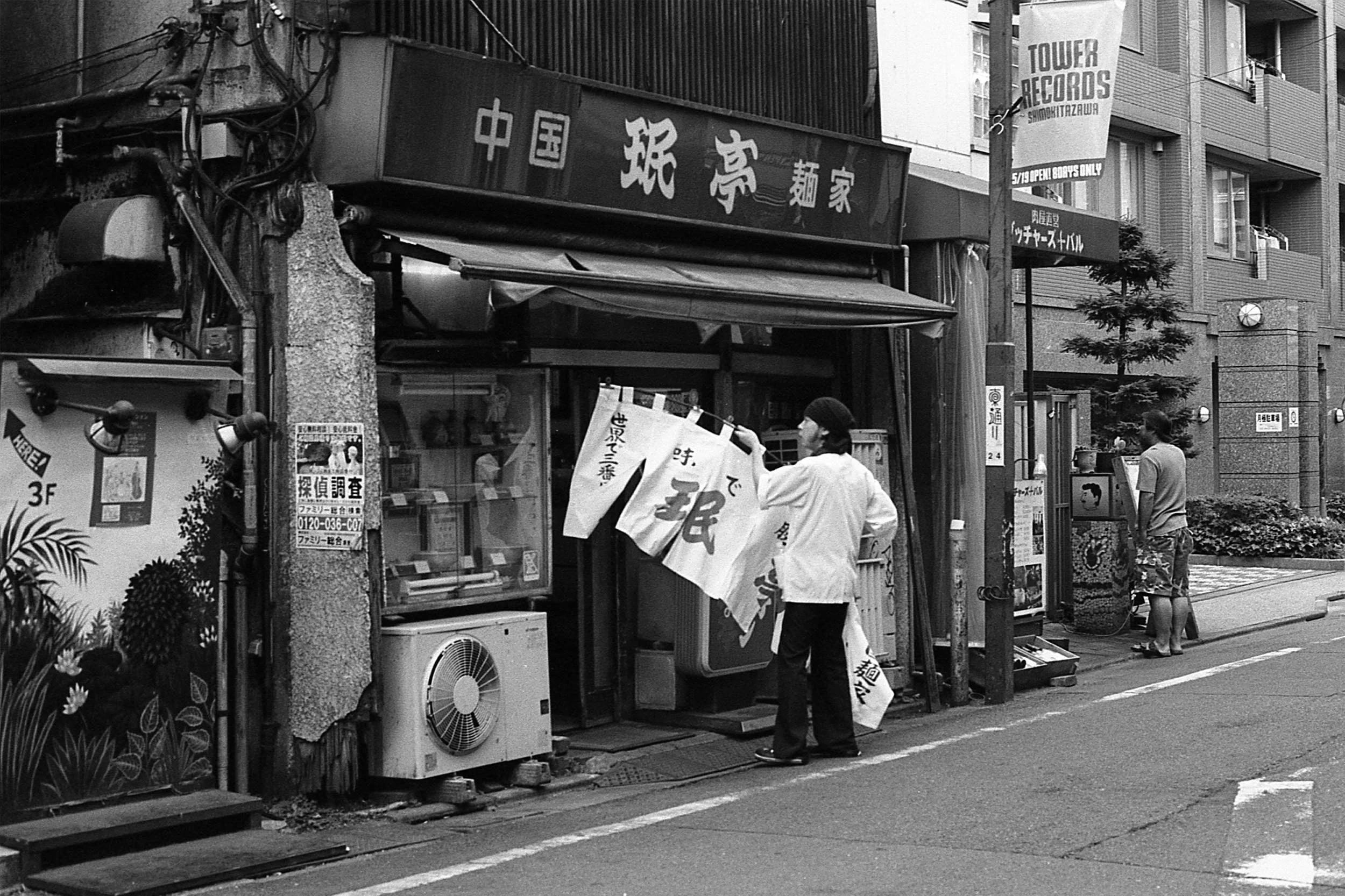 Black and white street scene featuring a small storefront with Japanese signs and a person hanging a banner outside, next to another person standing on the sidewalk. There are buildings, posters, a flag, and trees visible in the background.
