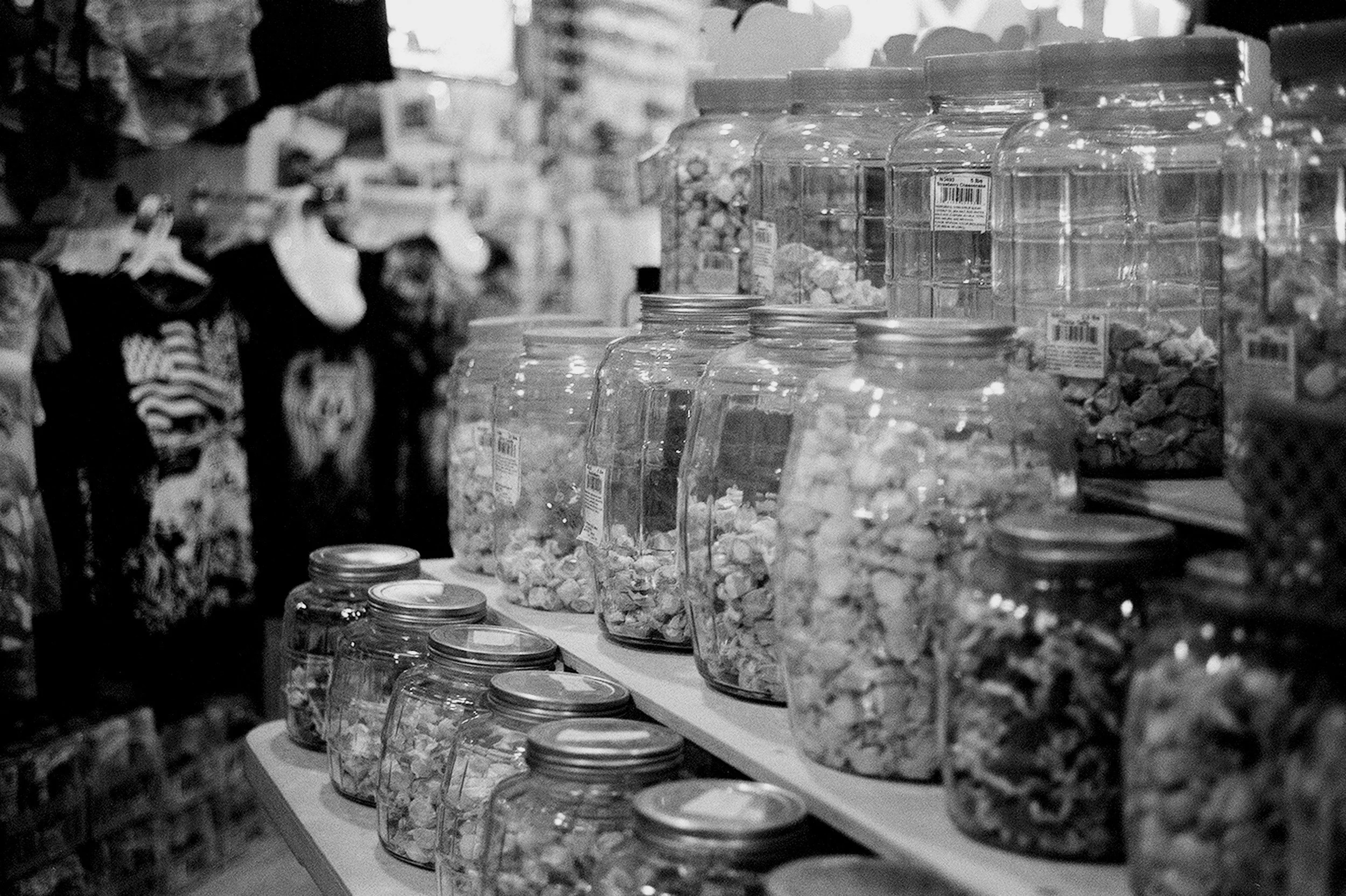 A black and white photo of a candy or snack aisle in a store, with jars and containers filled with various candies or small snacks arranged on shelves, and blurred figures of shoppers in the background.