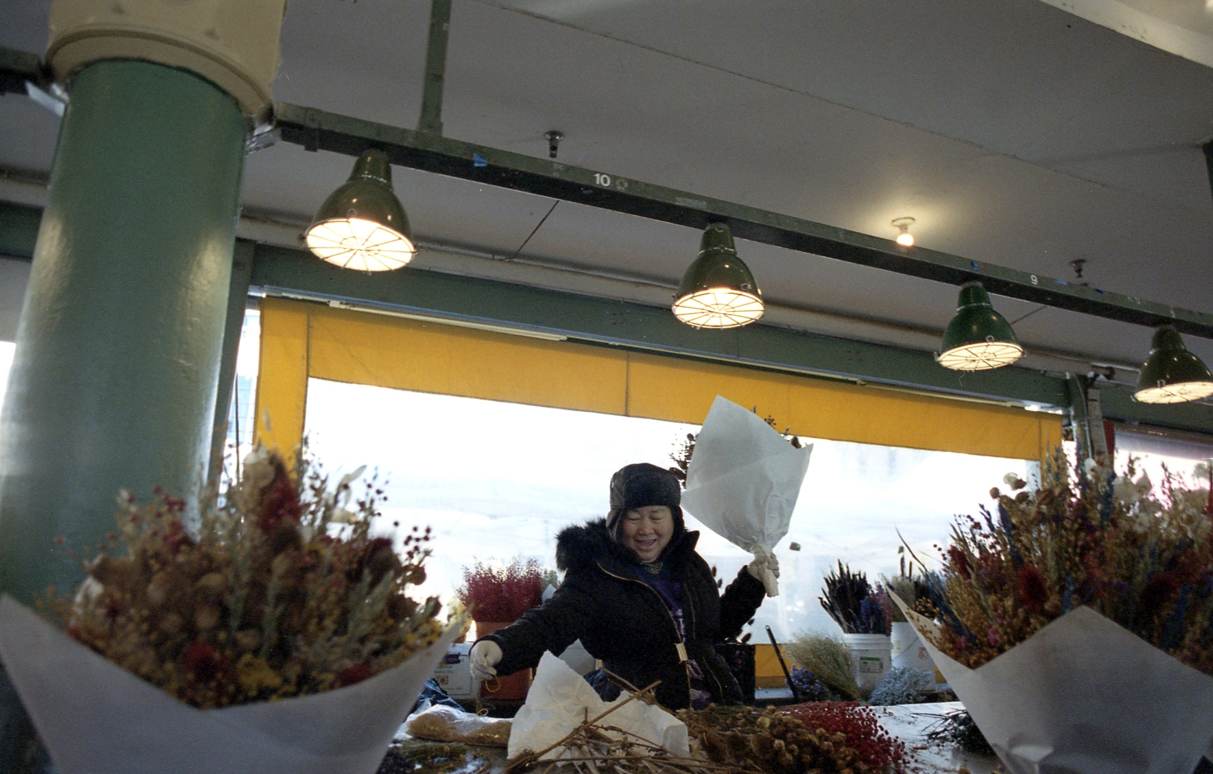 Woman at a flower shop working with flower bouquets at an outdoor market or event, with decorative lighting and a yellow awning visible in the background.