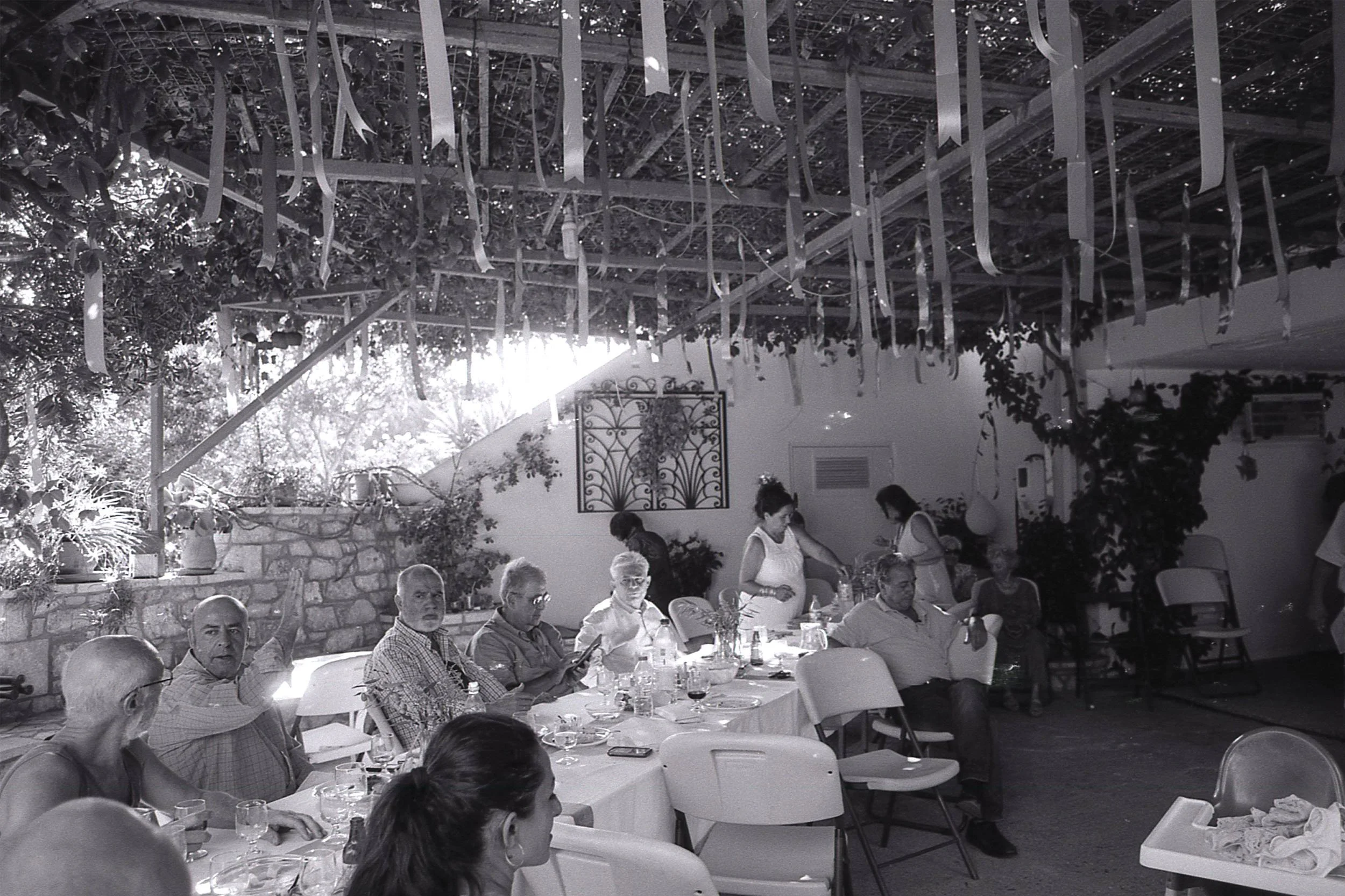 People attending a gathering or celebration in an outdoor patio with a wooden pergola overhead, decorated with ribbons and string lights, seated around tables with wine glasses.