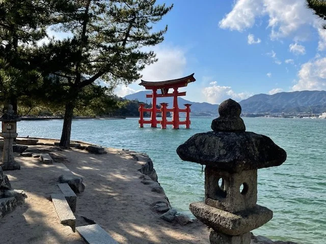 Scenic view of a large red torii gate in water, with a shoreline and trees in the foreground, mountains in the background, and a partly cloudy blue sky.