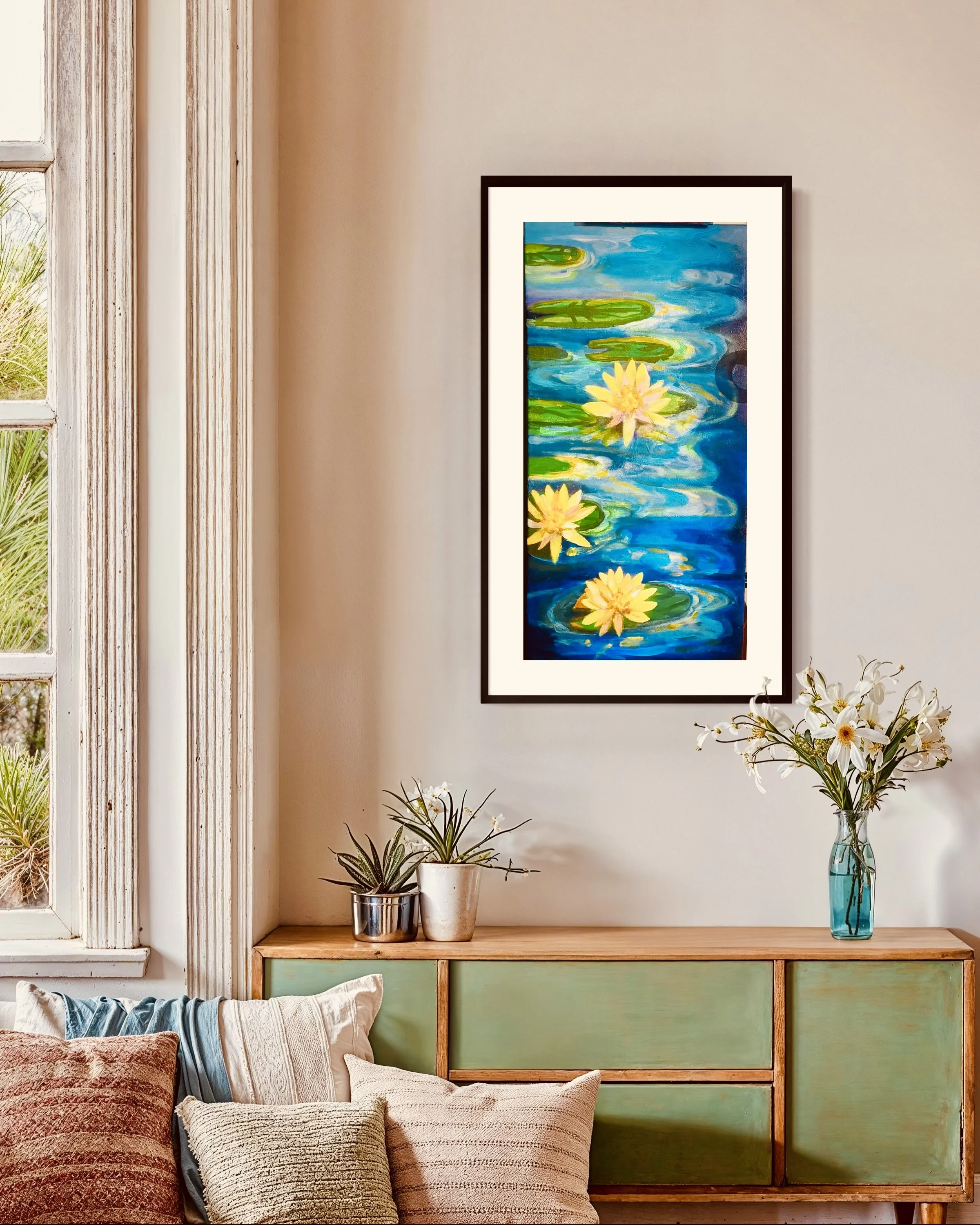 Interior of a living room with a framed painting of yellow water lilies on a pond hanging on a beige wall. Below the painting is a wooden sideboard with green drawers, decorated with potted plants and a tall glass vase of white flowers. Cushions are 