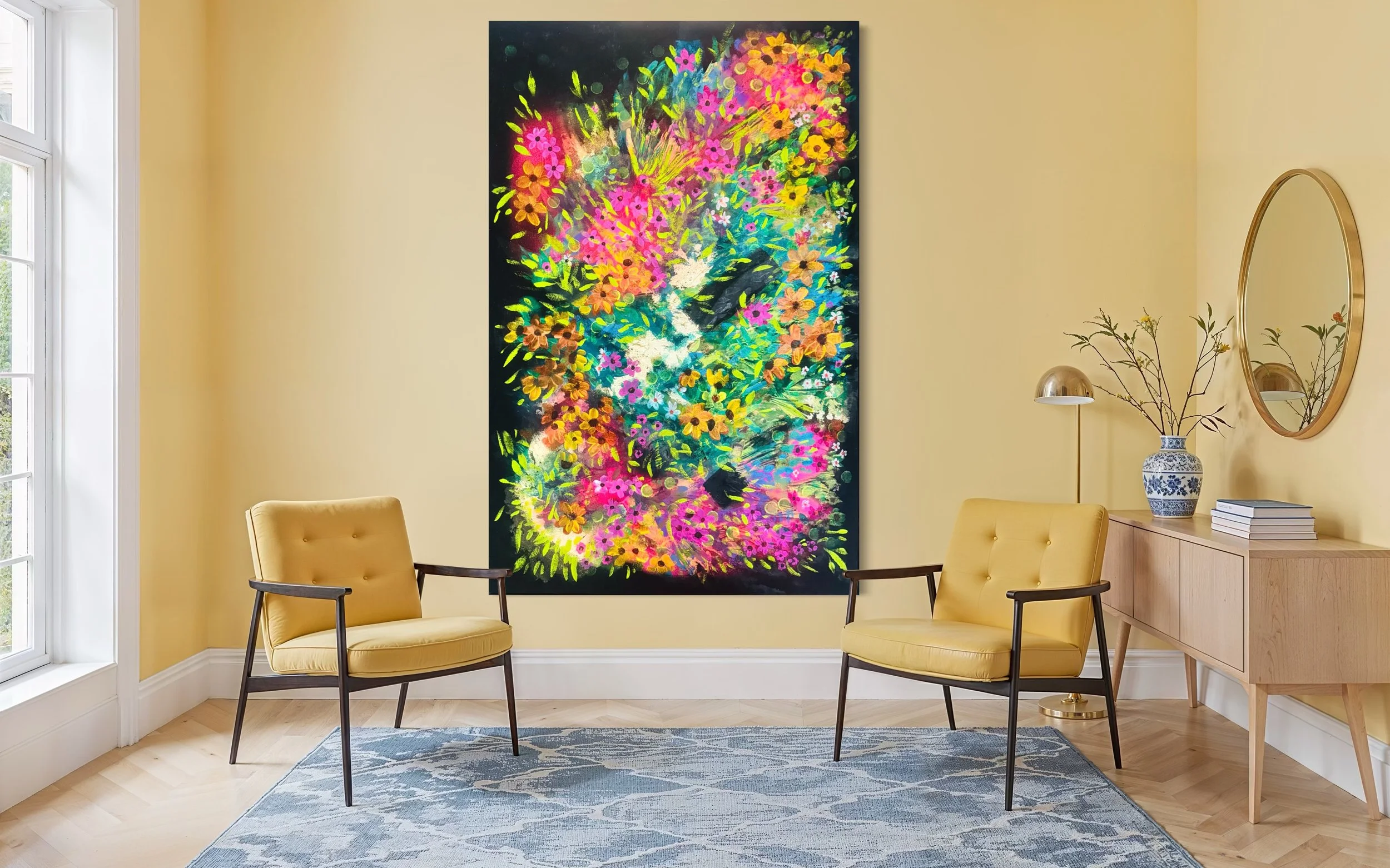 Living room with yellow walls, a large colorful floral painting, two yellow armchairs, a wooden sideboard with a vase of branches and a mirror above, and a window to the left.