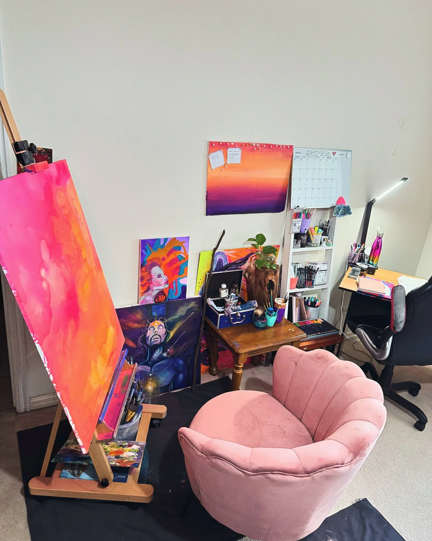 Art studio with a pink velvet armchair in front of a desk, multiple colorful paintings leaning against the wall, a small table with art supplies, and a whiteboard calendar.