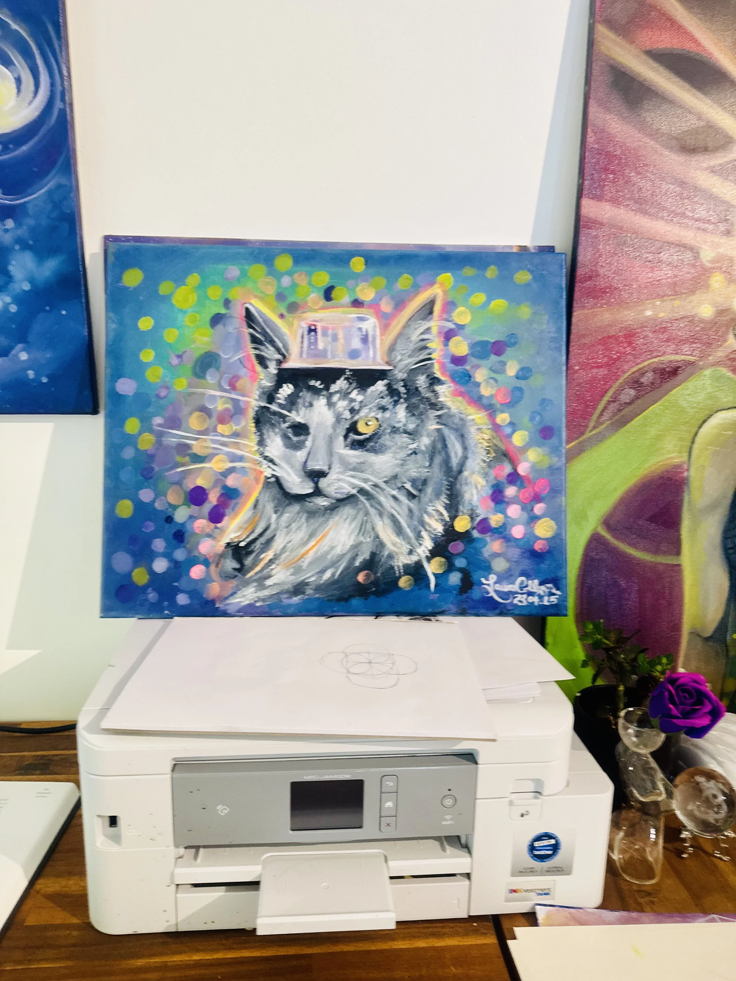Colorful painting of a cat wearing a hat, surrounded by vibrant dots, on a desk with printing equipment.