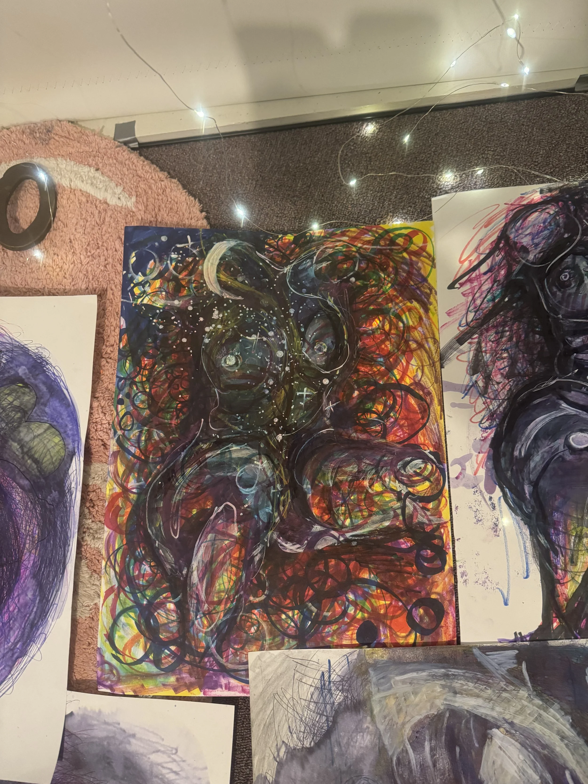 Colorful abstract painting of a figure, with dark outlines and swirling patterns, on a background of vibrant reds, oranges, and other colors, displayed on a floor next to other artworks and fairy lights.