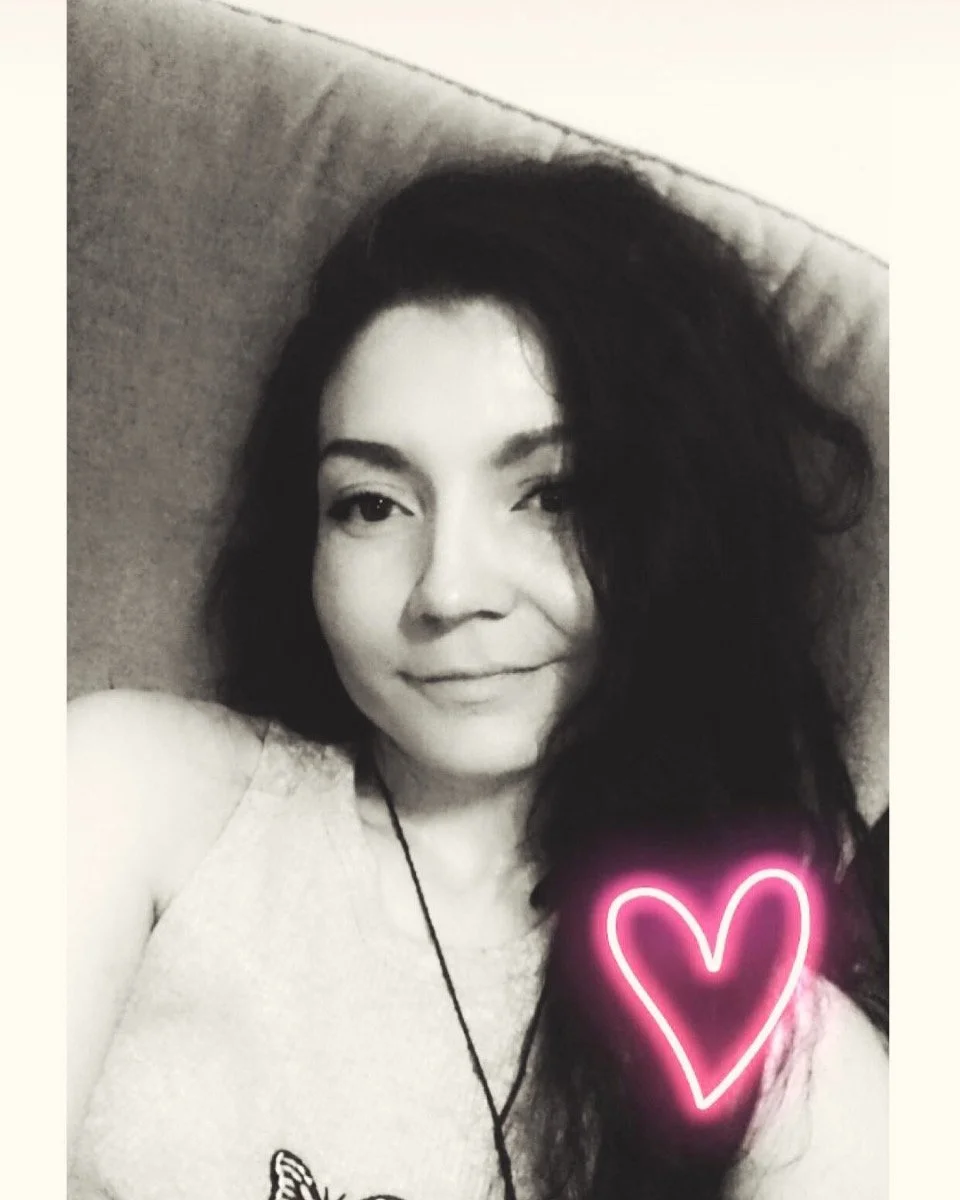 A woman with dark, wavy hair lying on a bed, smiling, in black and white with a pink neon heart overlay near her shoulder.