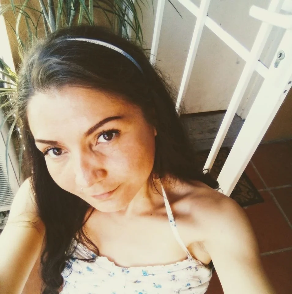 A young woman with long dark hair, wearing a floral spaghetti strap top, taking a selfie indoors with a plant and a white structure in the background.